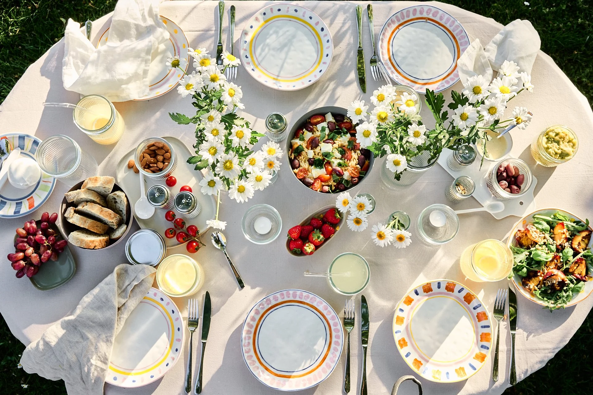 Ein gedeckter Esstisch im Freien mit weißen Tellern, Besteck, Gläsern, Blumen und verschiedenen Speisen wie Salat, Früchte, Brot, Getränke und Snacks.