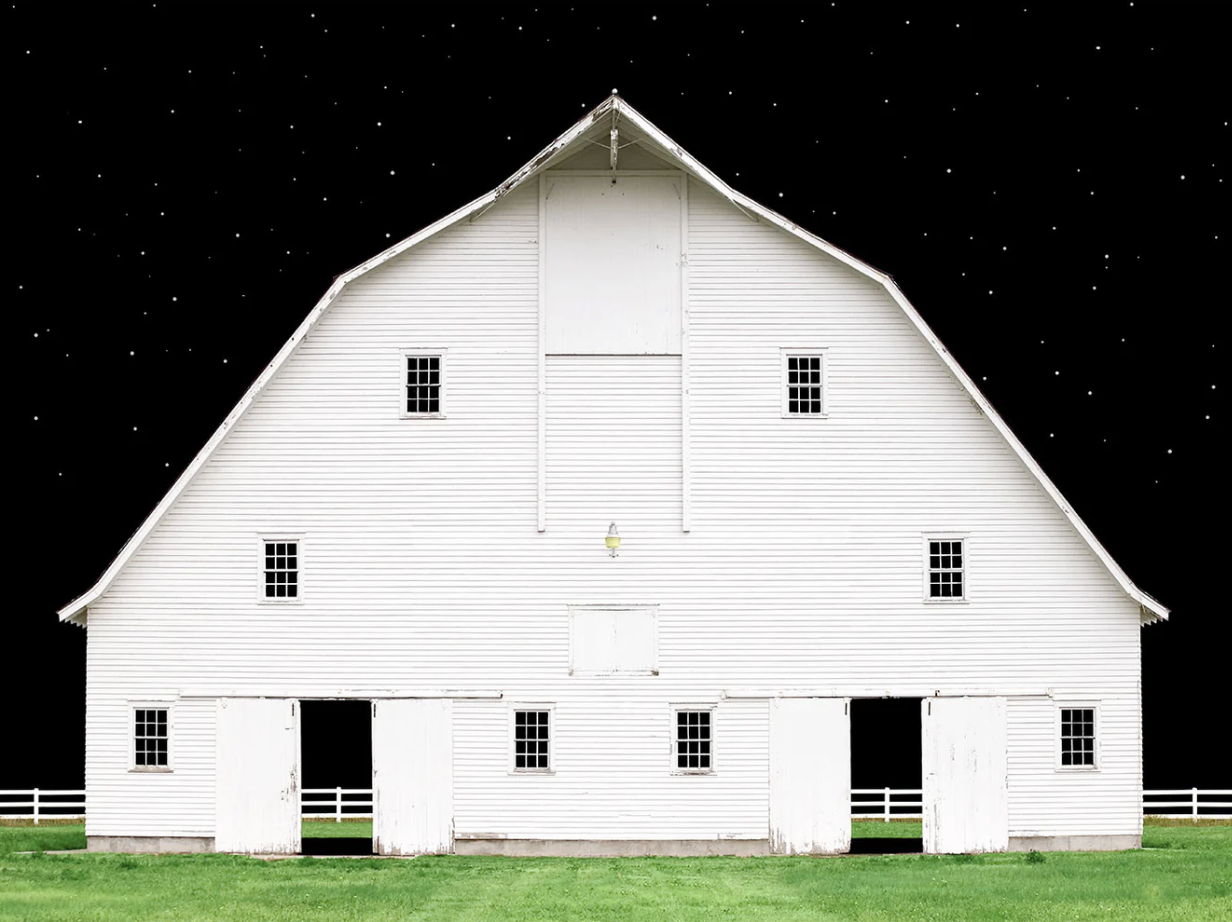 A large white barn with multiple small windows and open doors, situated on a grassy field under a night sky with stars.