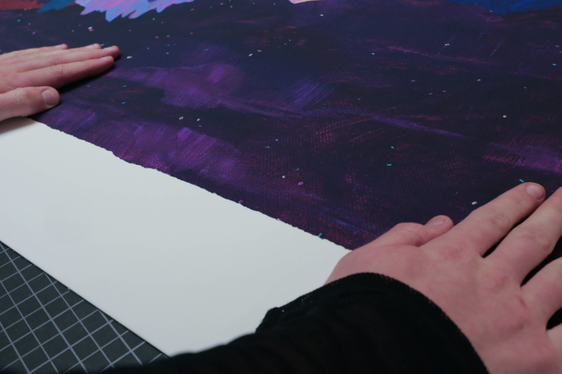 Hands holding and stretching a dark purple and black painted canvas with small specks of light blue and pink, on a table with a cutting mat.