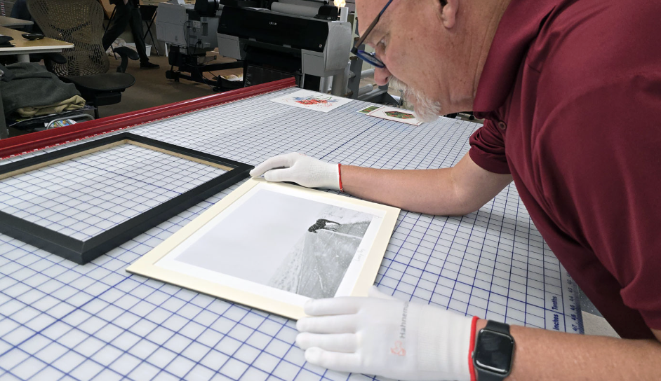 Person carefully handling a framed black-and-white photograph on a large cutting mat in a workspace or archive setting.
