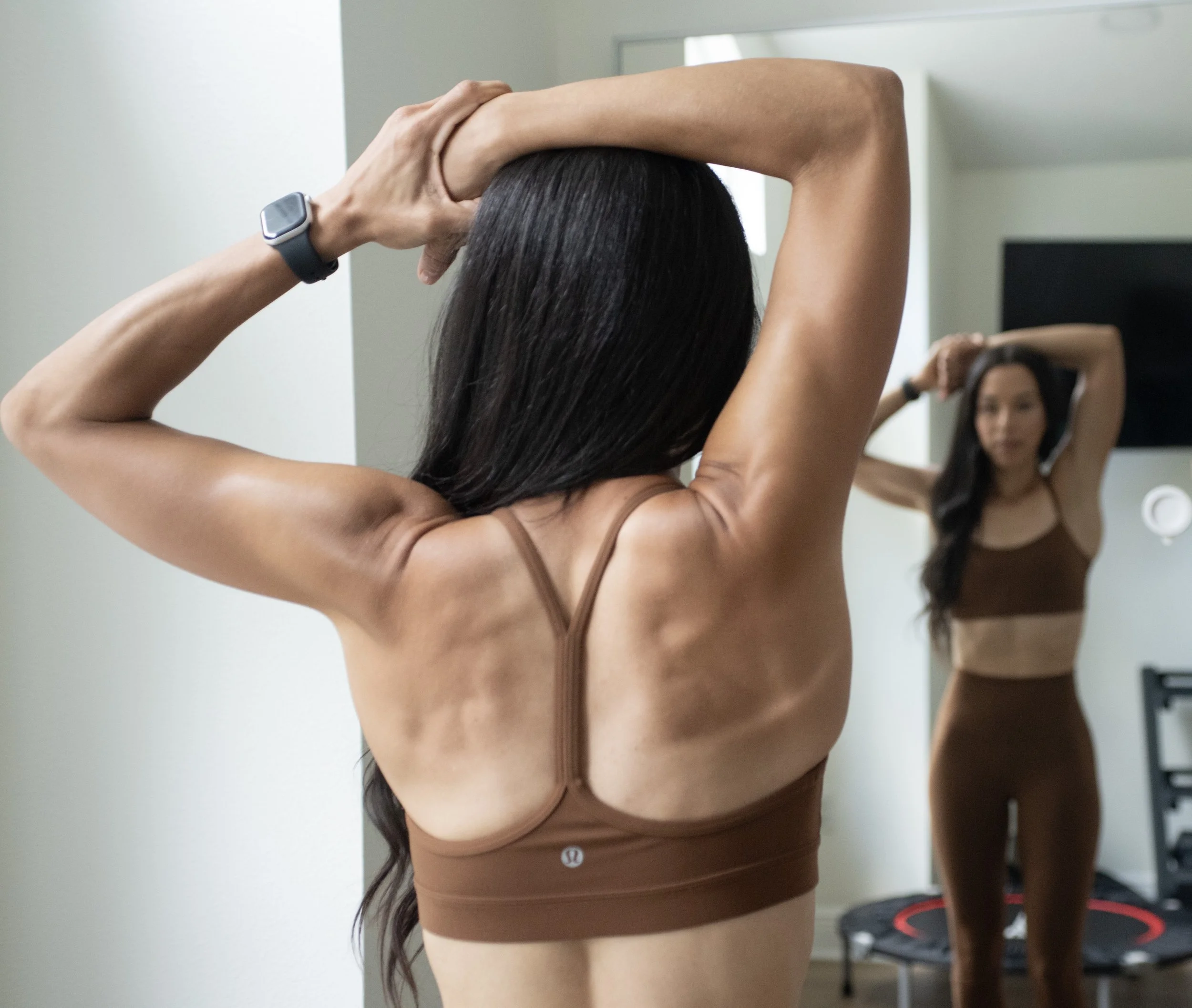 A woman with long dark hair tying her hair in front of a mirror, wearing a brown sports bra and matching shorts, with an Apple Watch on her left wrist. The background includes a white wall, a door, and a small trash bin with a black and red lid.