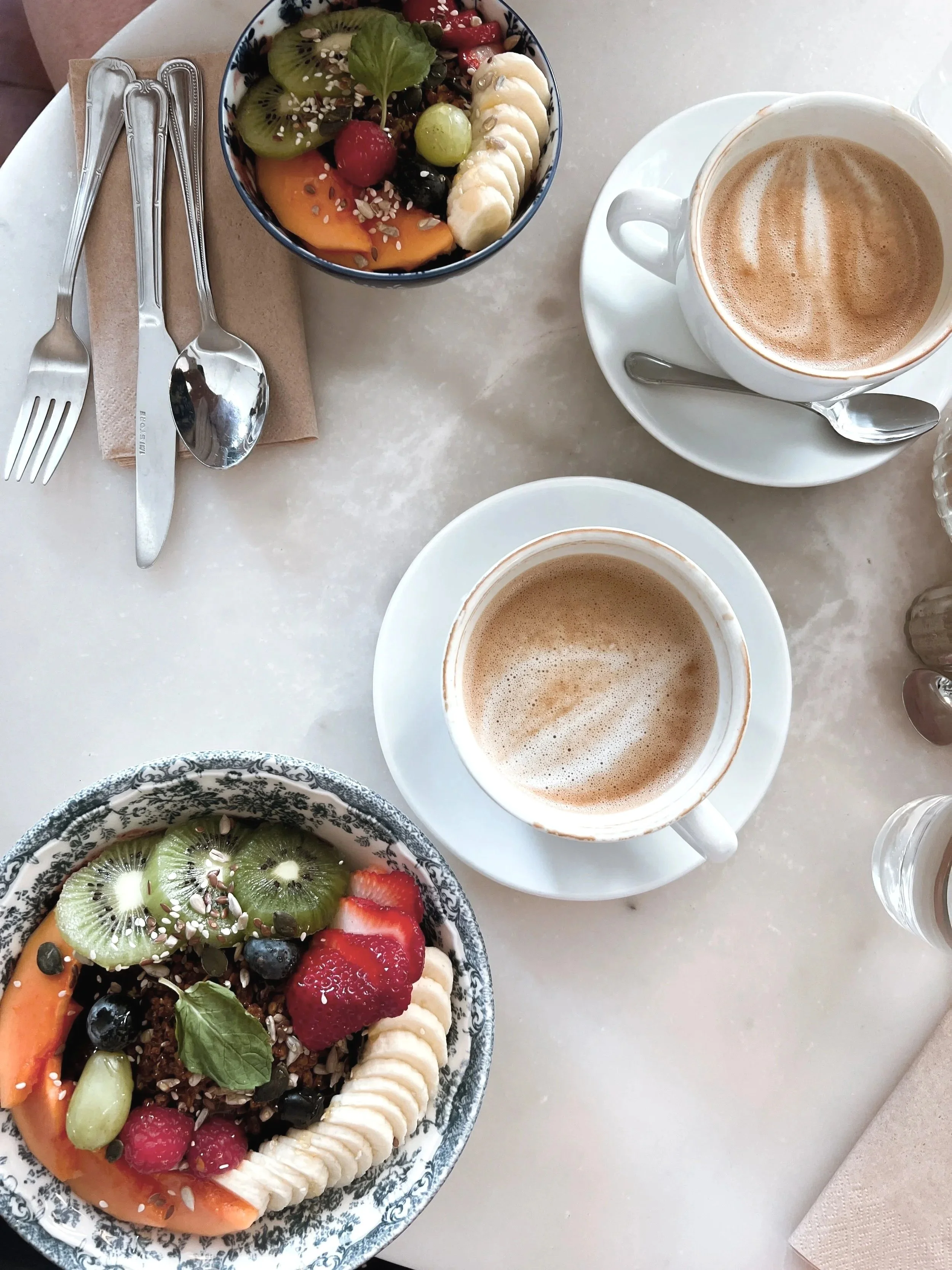 Two cups of coffee with frothy whipped topping on a white table next to a bowl of mixed fresh fruit and granola, including sliced banana, kiwi, strawberries, blueberries, grapes, and a slice of peach, with utensils and napkins nearby.