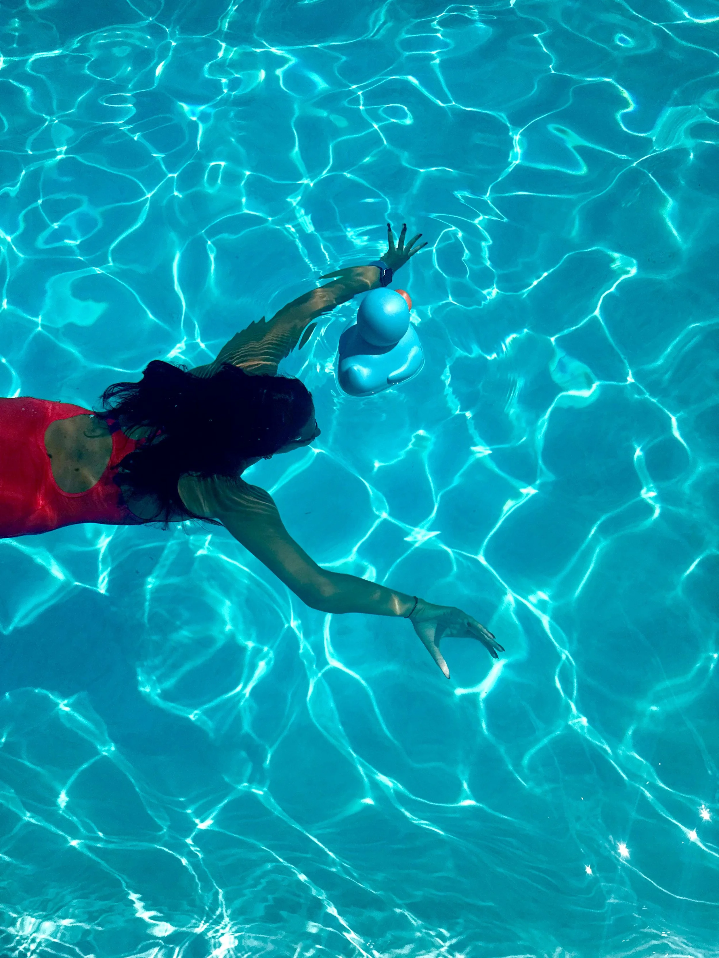 A woman in a red swimsuit floating in a swimming pool, reaching towards a rubber duck in the water.