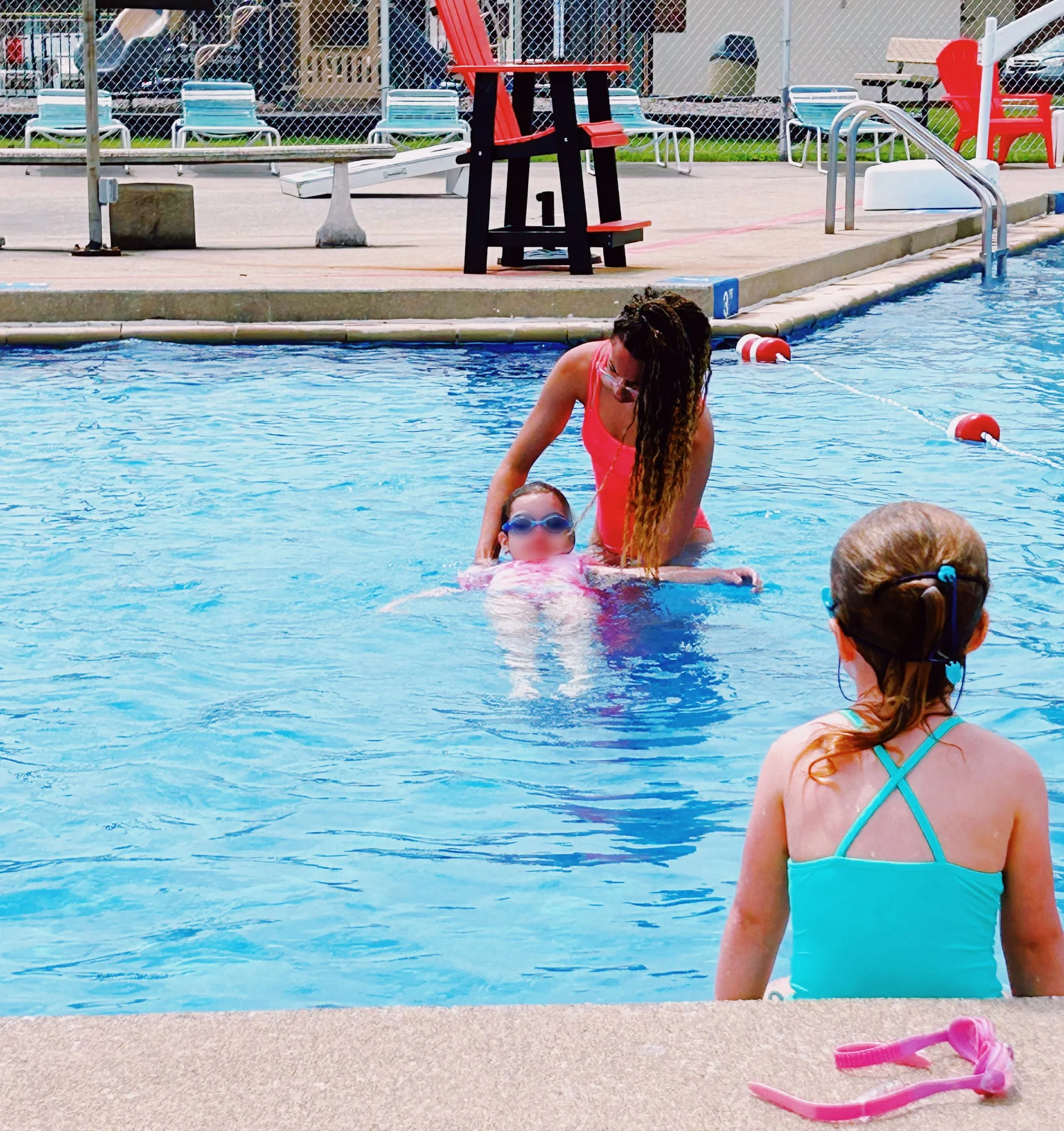 A swimming pool scene with a woman teaching two young girls how to swim. The woman is in the water supporting one girl, who is wearing goggles, while the other girl sits on the pool edge wearing a swimsuit and goggles. Pool toys and chairs are visibl