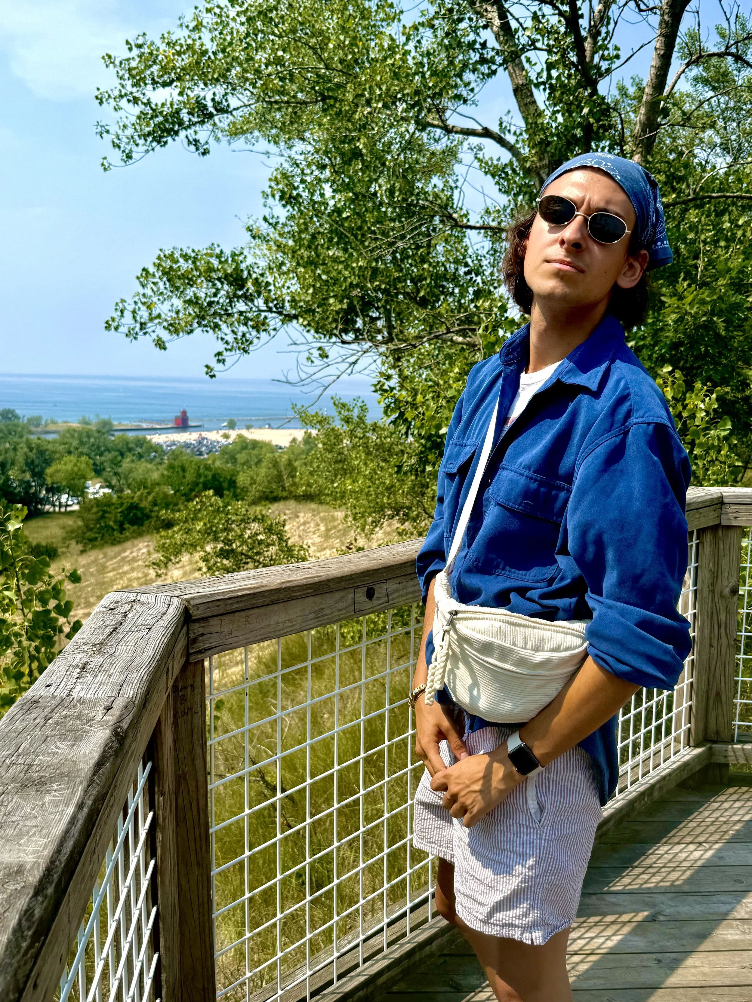 A young man wearing sunglasses, a blue bandana, a blue jacket, a white fanny pack, and striped shorts standing on a wooden observation deck with trees and a beach with a lighthouse in the background.