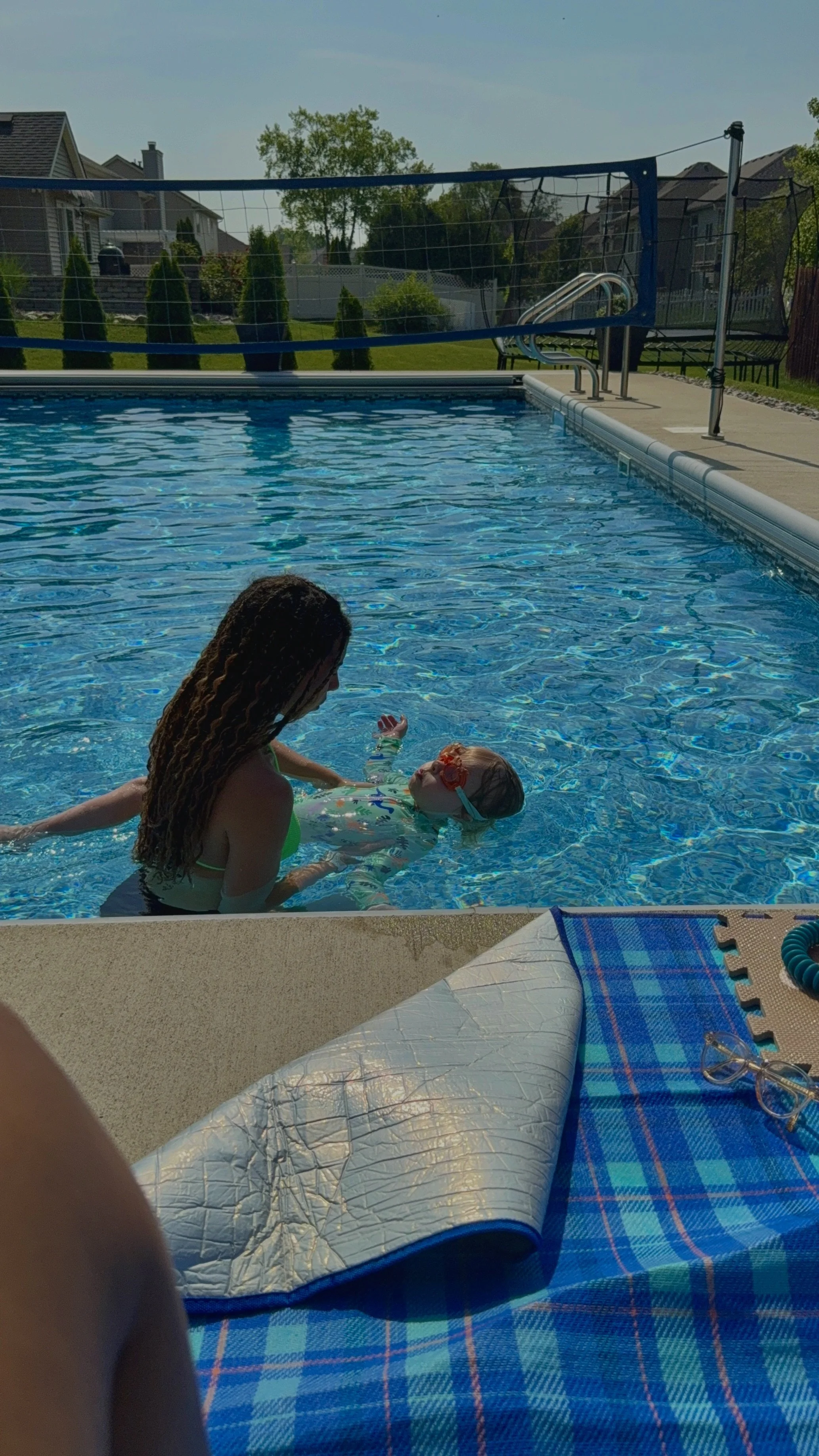 A woman helping a young girl swimming in a backyard pool on a sunny day.