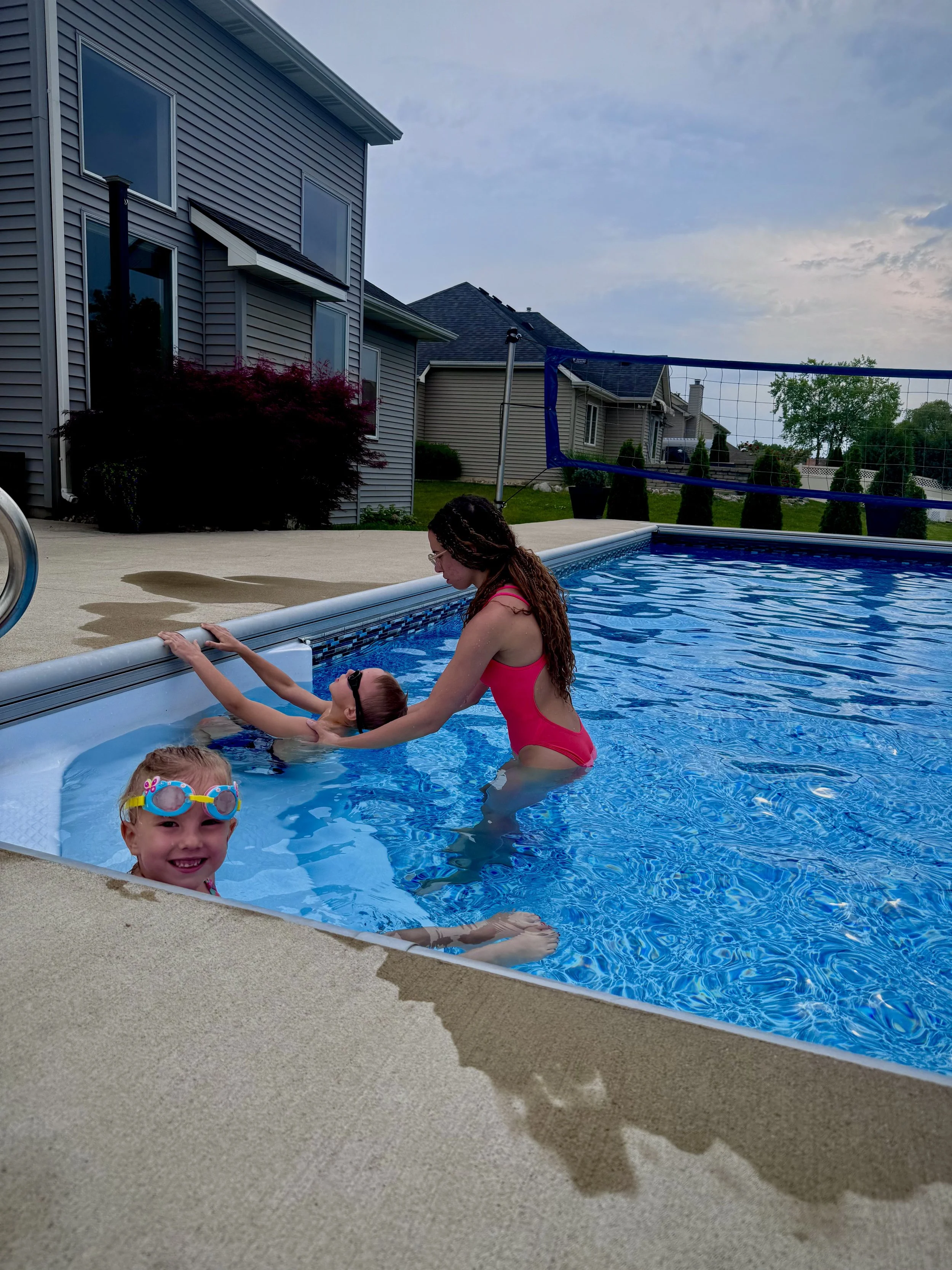 A woman helps a young girl exit a swimming pool while a smiling girl with goggles swims nearby, in a backyard with houses and a volleyball net visible in the background.