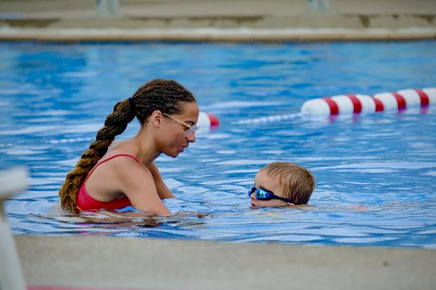 A young woman with braided hair and glasses assisting a young boy with goggles in a swimming pool.