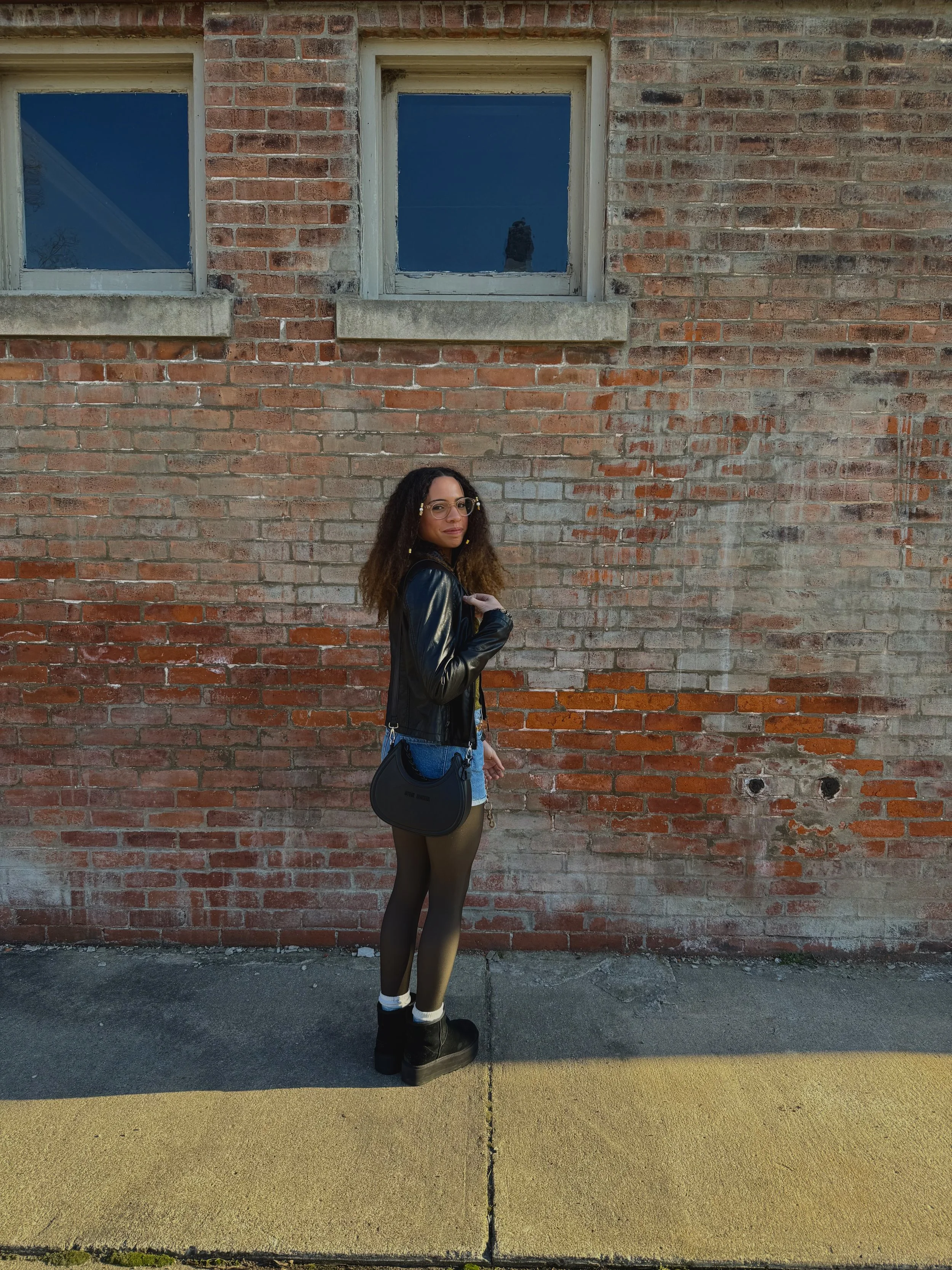 A woman standing on a sidewalk against a brick wall, wearing glasses, a black leather jacket, denim shorts, black tights, black boots, and carrying a black purse.