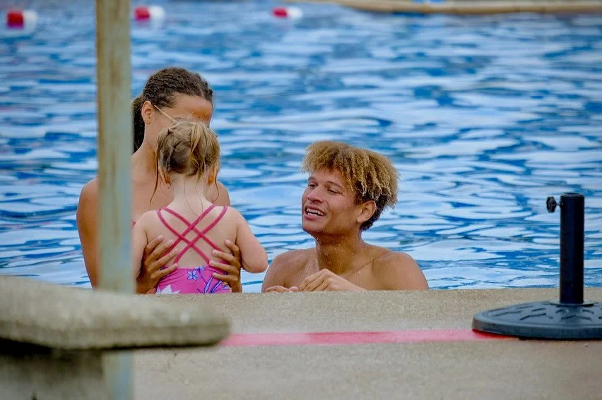 A young man in a swimming pool holding a small girl in a pink swimsuit, with a woman standing nearby, all smiling.