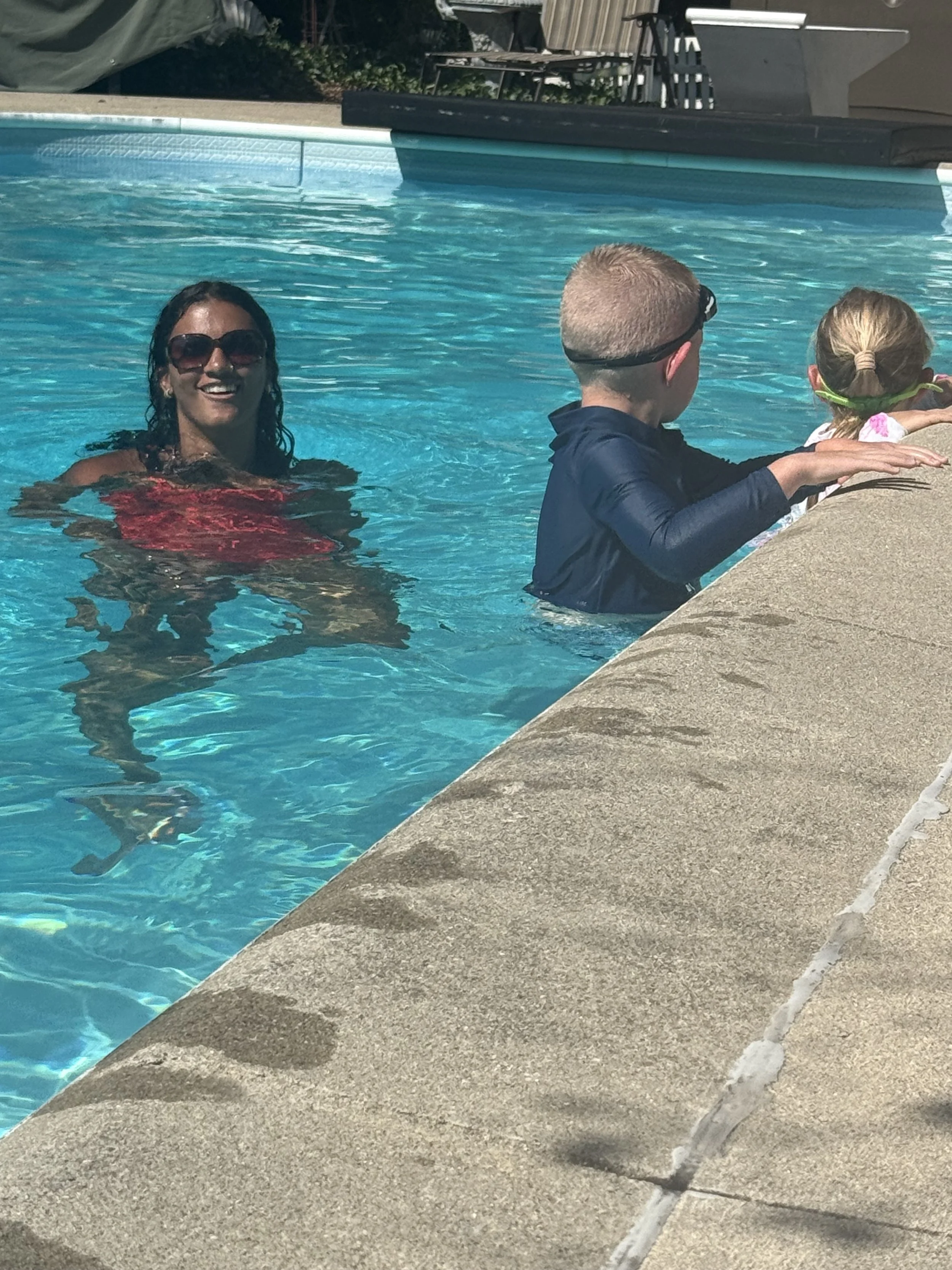 A woman in sunglasses and a red swimsuit smiling in a swimming pool with two children, one boy and one girl, near the pool's edge.