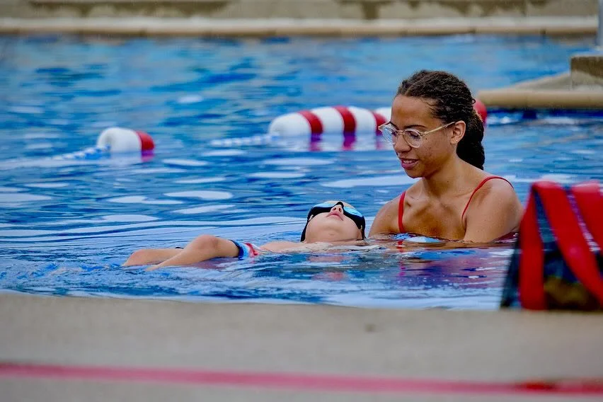 A woman and a young boy in a swimming pool, with the woman helping the boy float on his back, wearing goggles, near pool lane dividers.