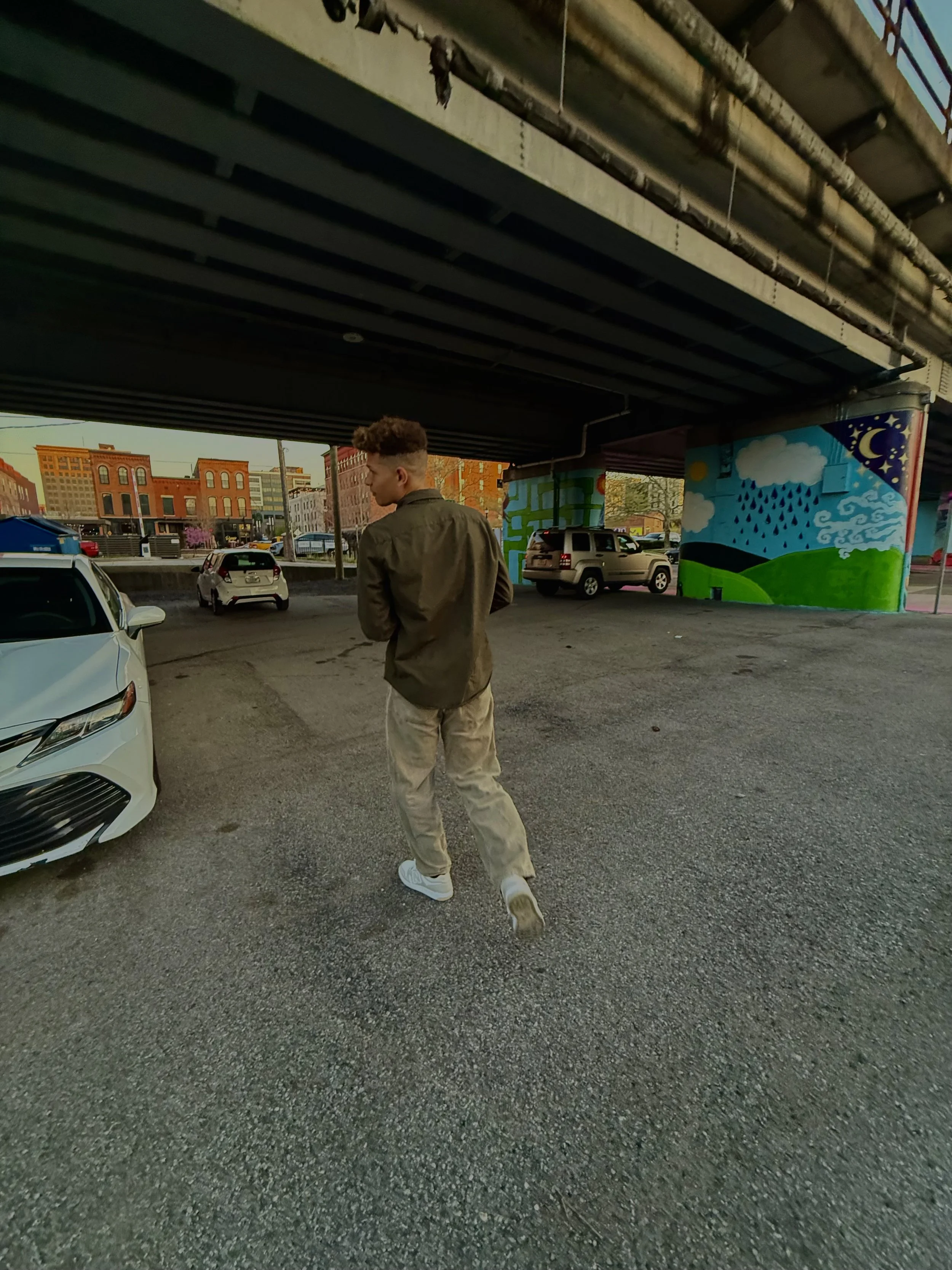 Young man walking away under an overpass, with colorful streetside murals, parked cars, and urban buildings in the background.