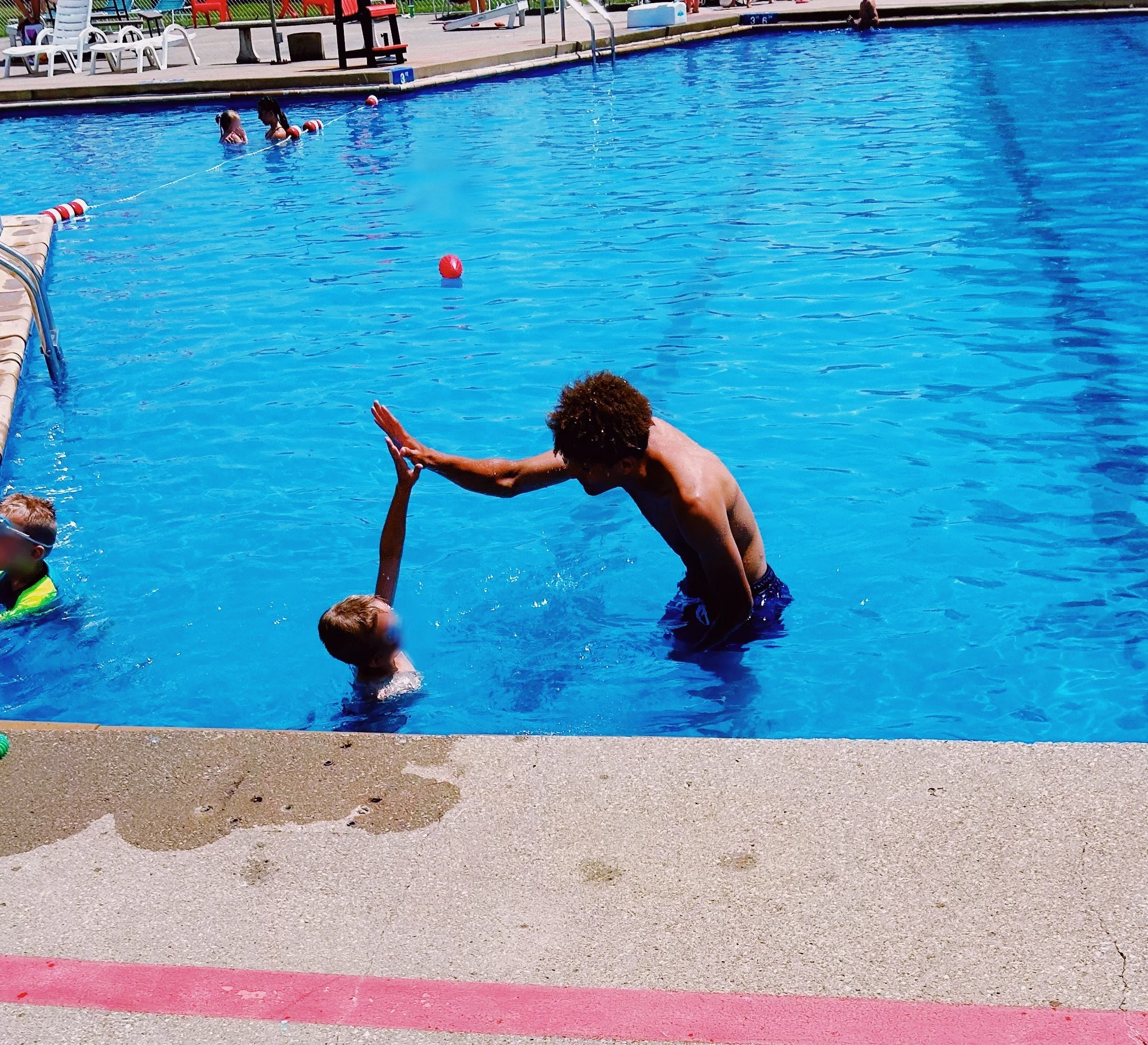 A boy giving a high five to a girl in a swimming pool, with other children swimming and playing in the background.