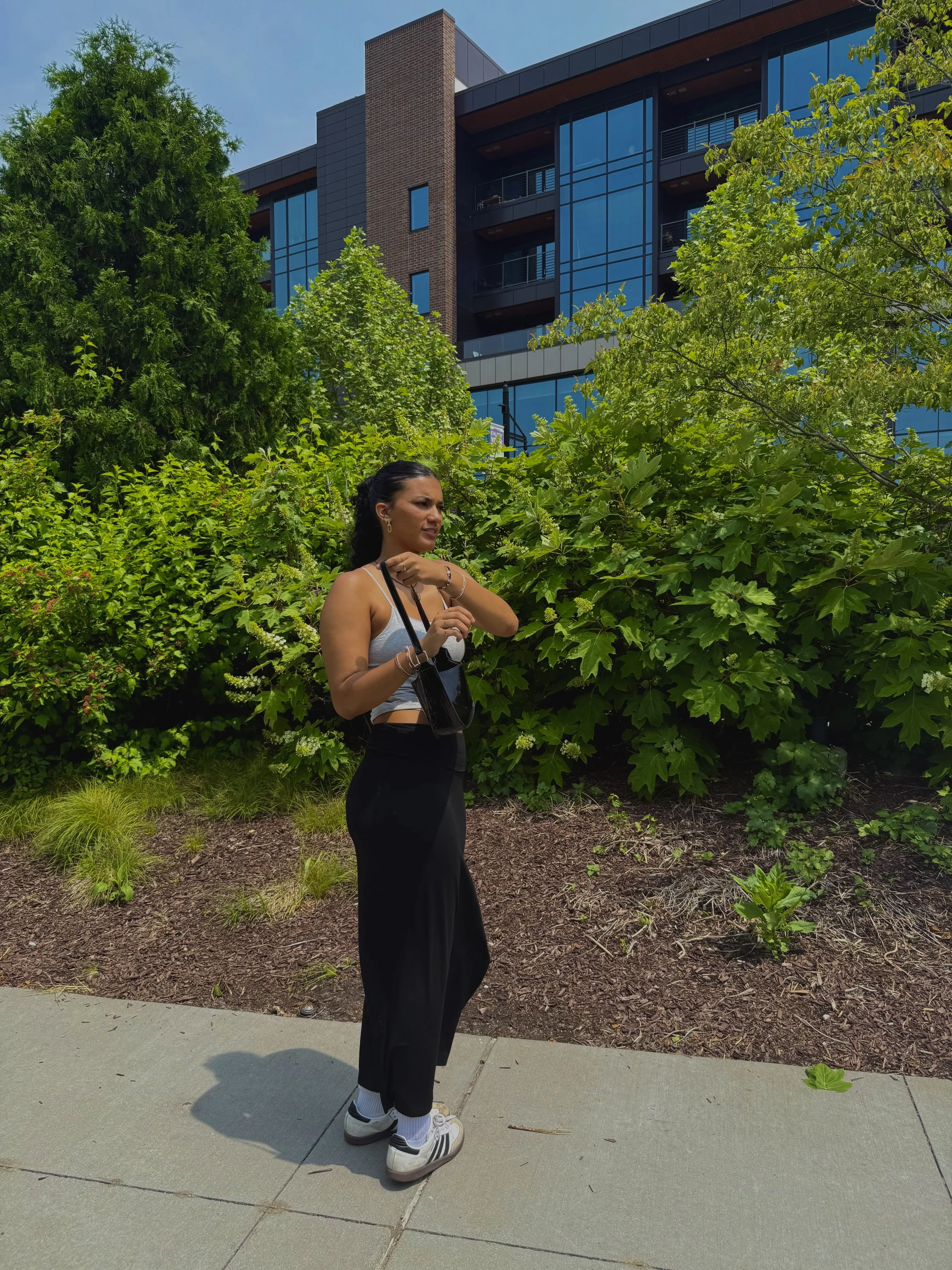 A woman standing on a sidewalk next to green bushes, with modern glass apartment buildings in the background. She is wearing a gray tank top, black pants, and sneakers, carrying a small black purse, and looking to her left.