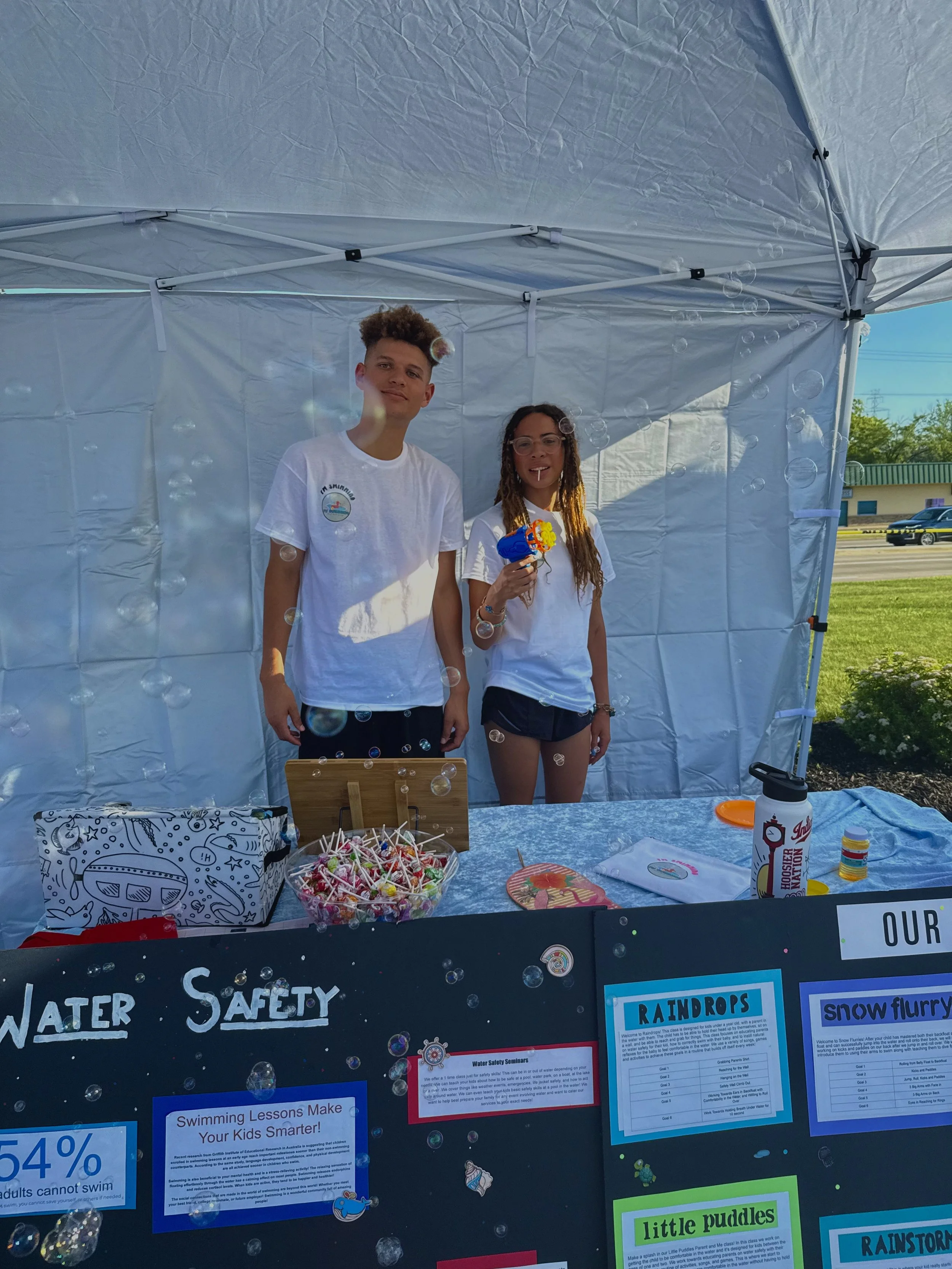 Two teenagers standing under a white canopy tent, with water safety and swimming promotional posters on a blackboard in front of them, surrounded by small bubbles, with a grassy area and parked cars in the background.