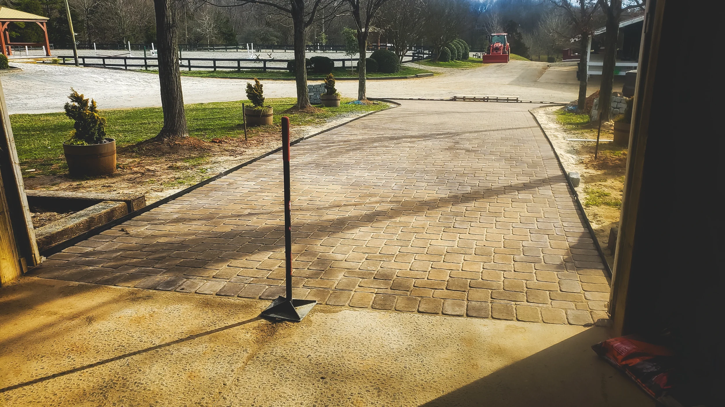 View from inside a building looking out onto a newly paved brick driveway with a construction barrier in the foreground. In the background, there are trees, potted plants, and a red tractor on a dirt road.