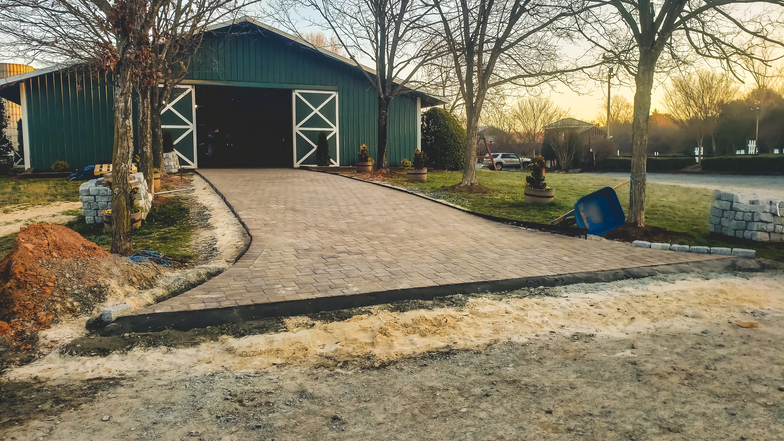 A newly paved driveway leading to a green barn with white trim in a yard during sunset, with trees, potted plants, and construction materials nearby.