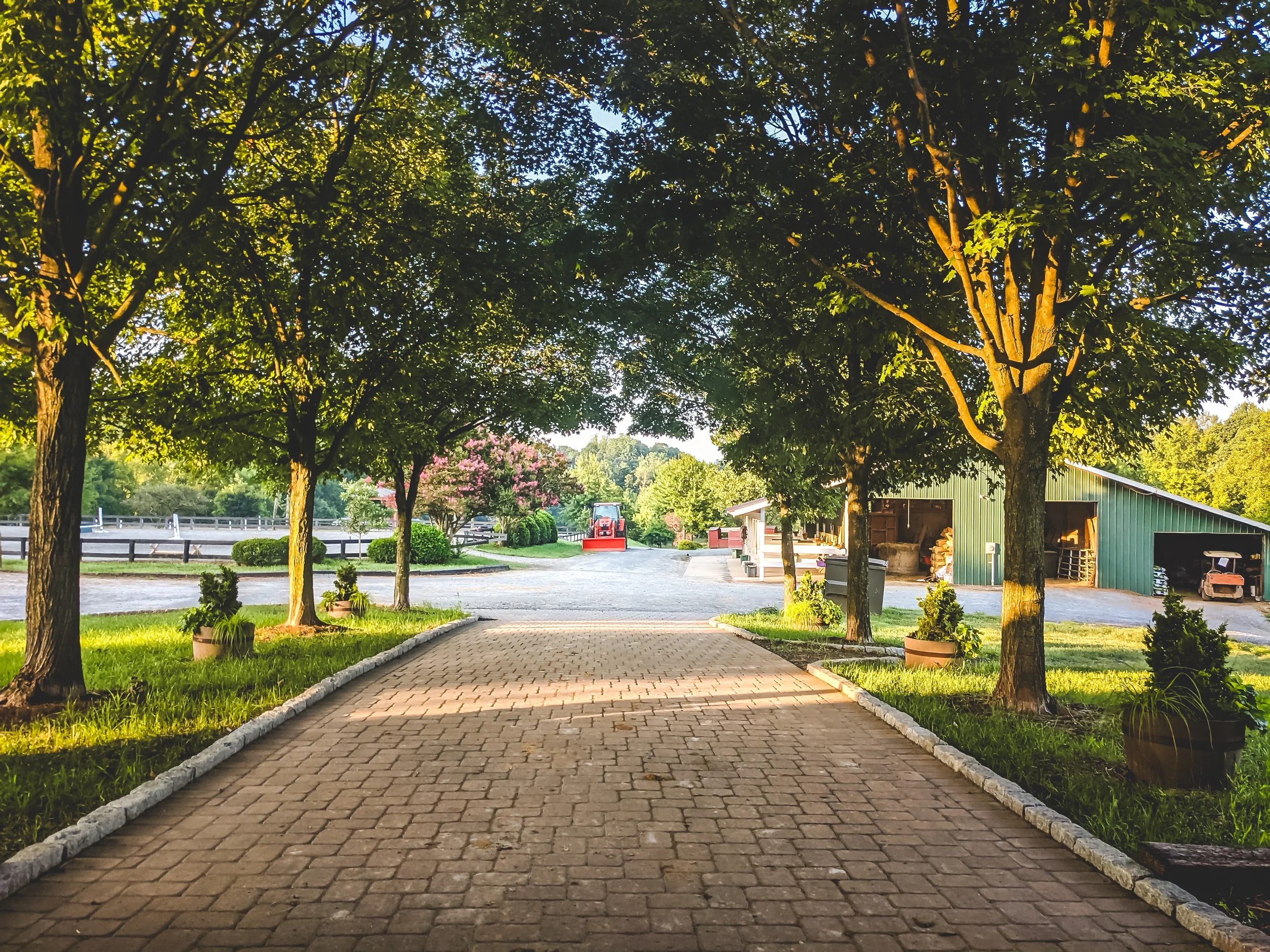 A paved brick pathway leading towards a farm scene with trees on either side. There is a green barn with an open door, and a red tractor in the background. Potted plants line the pathway and the area is bathed in warm sunlight.