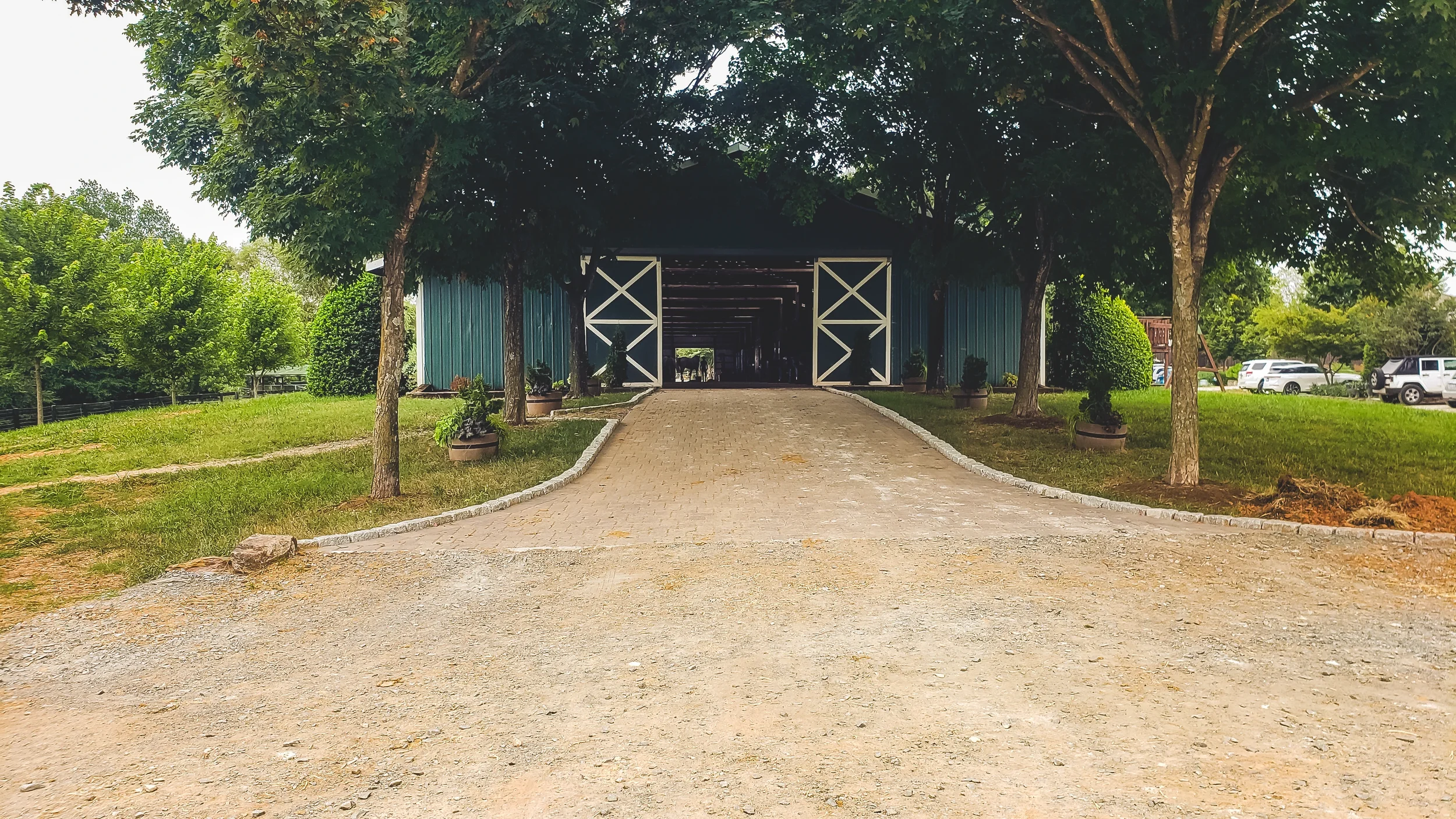 A covered green barn with white doors, surrounded by green trees and grass, with a paved driveway leading to the entrance.
