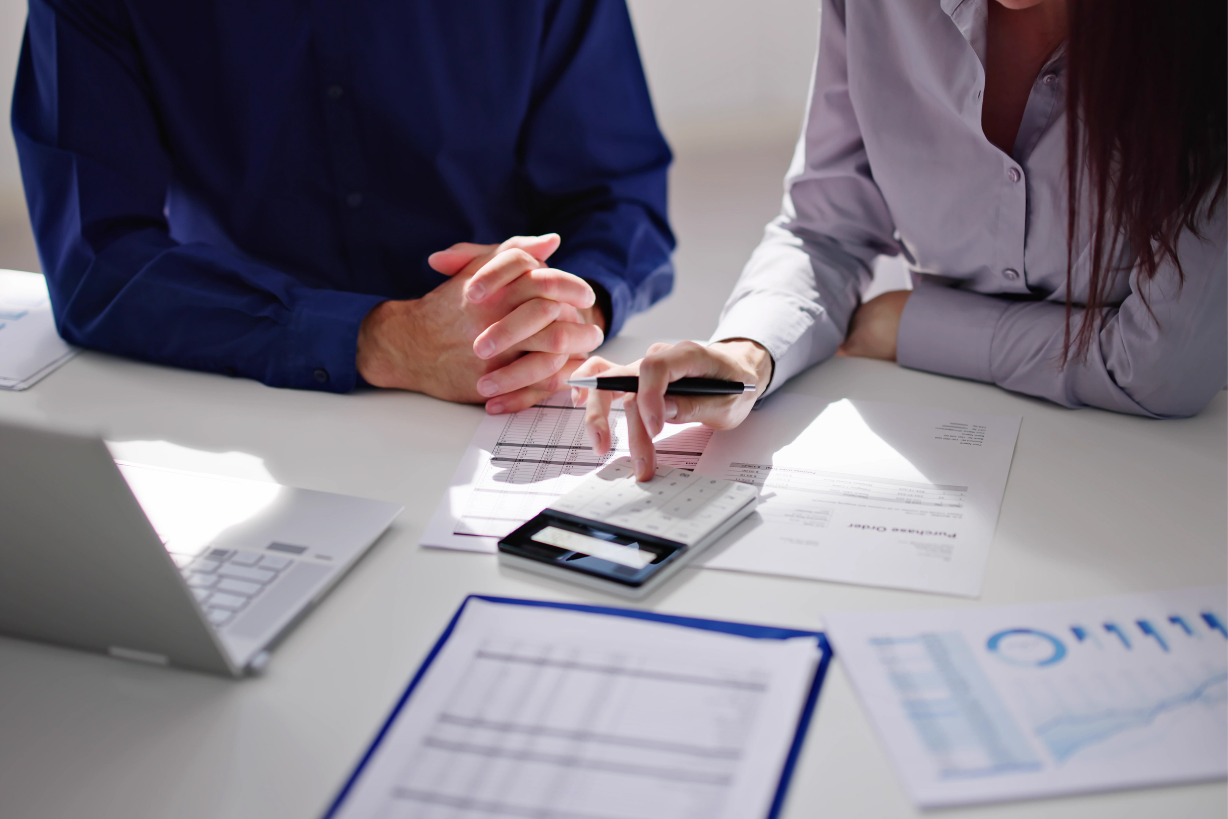 Two individuals at a desk reviewing financial documents, with one person pointing at a calculator, surrounded by papers and a laptop.
