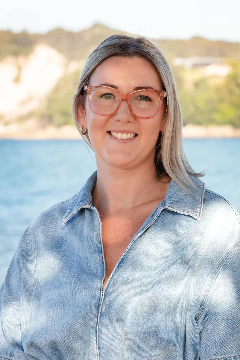 A woman with gray hair and pink glasses smiling outdoors near water with hills in the background.