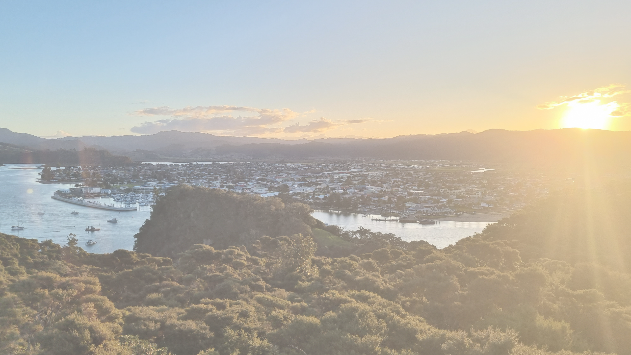 A scenic view of a river with boats, a town in the background, and a mountain range with the sun setting or rising, casting a warm glow over the landscape.