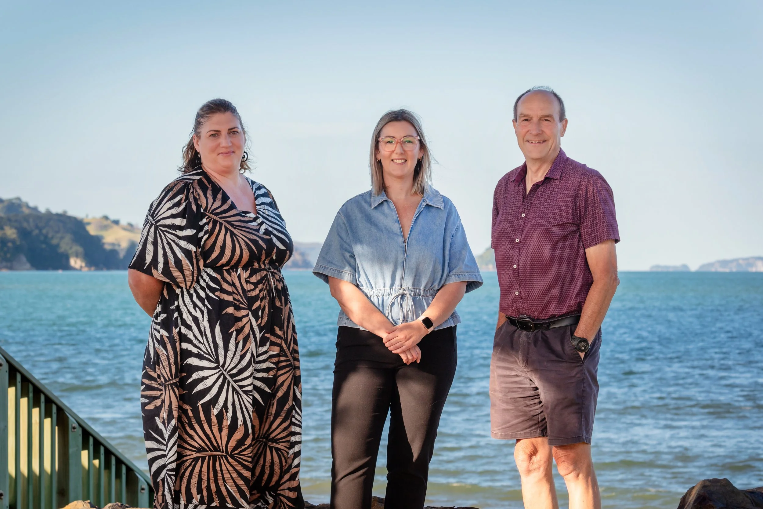 Three people standing on a shoreline near the water with distant hills, smiling at the camera on a sunny day.