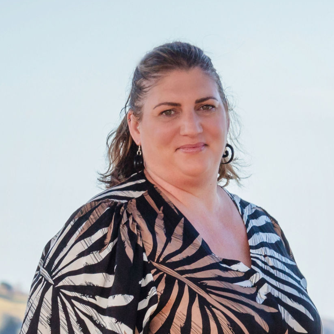 A woman with shoulder-length brown hair, wearing a black and white leaf-patterned blouse, and black earrings, standing outdoors with a plain light sky background.