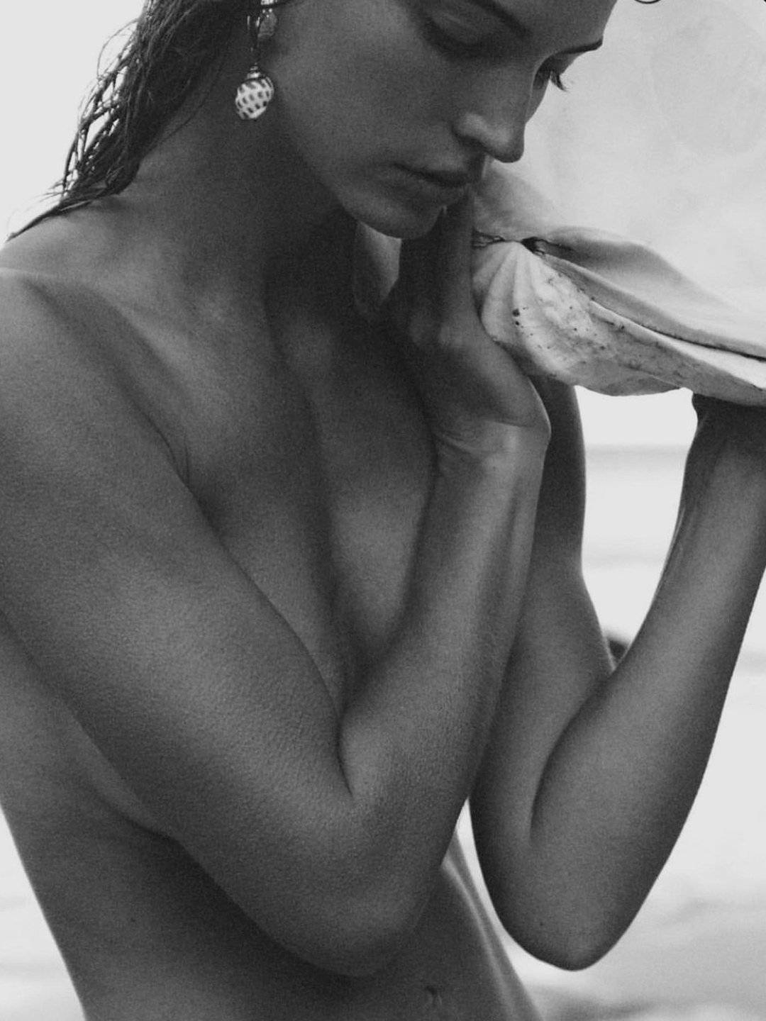 A black and white photograph of a woman holding a large seashell close to her face, wearing earrings, with wet hair and a contemplative expression.