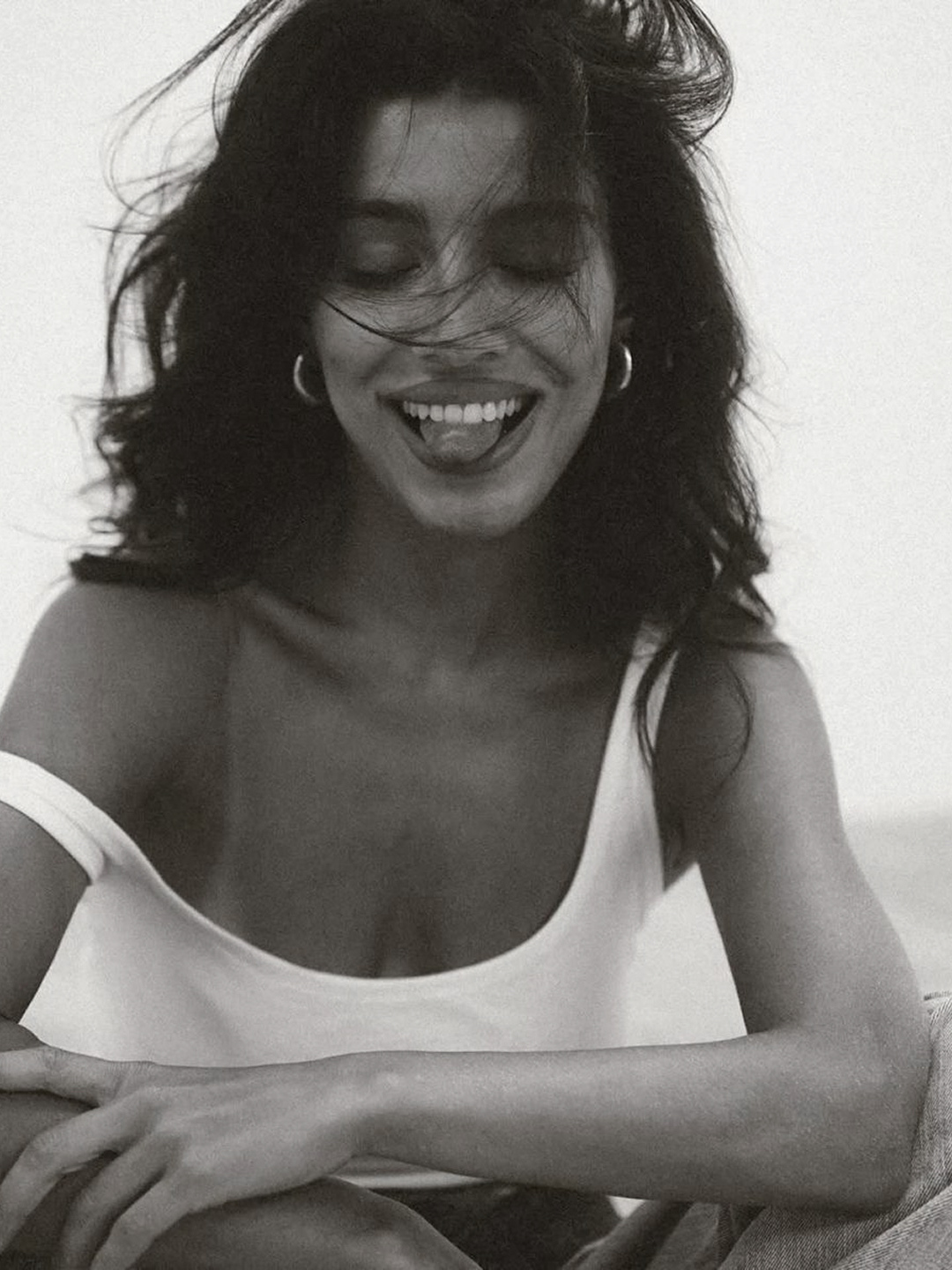 A woman with curly hair smiling and sticking her tongue out, wearing a sleeveless top.