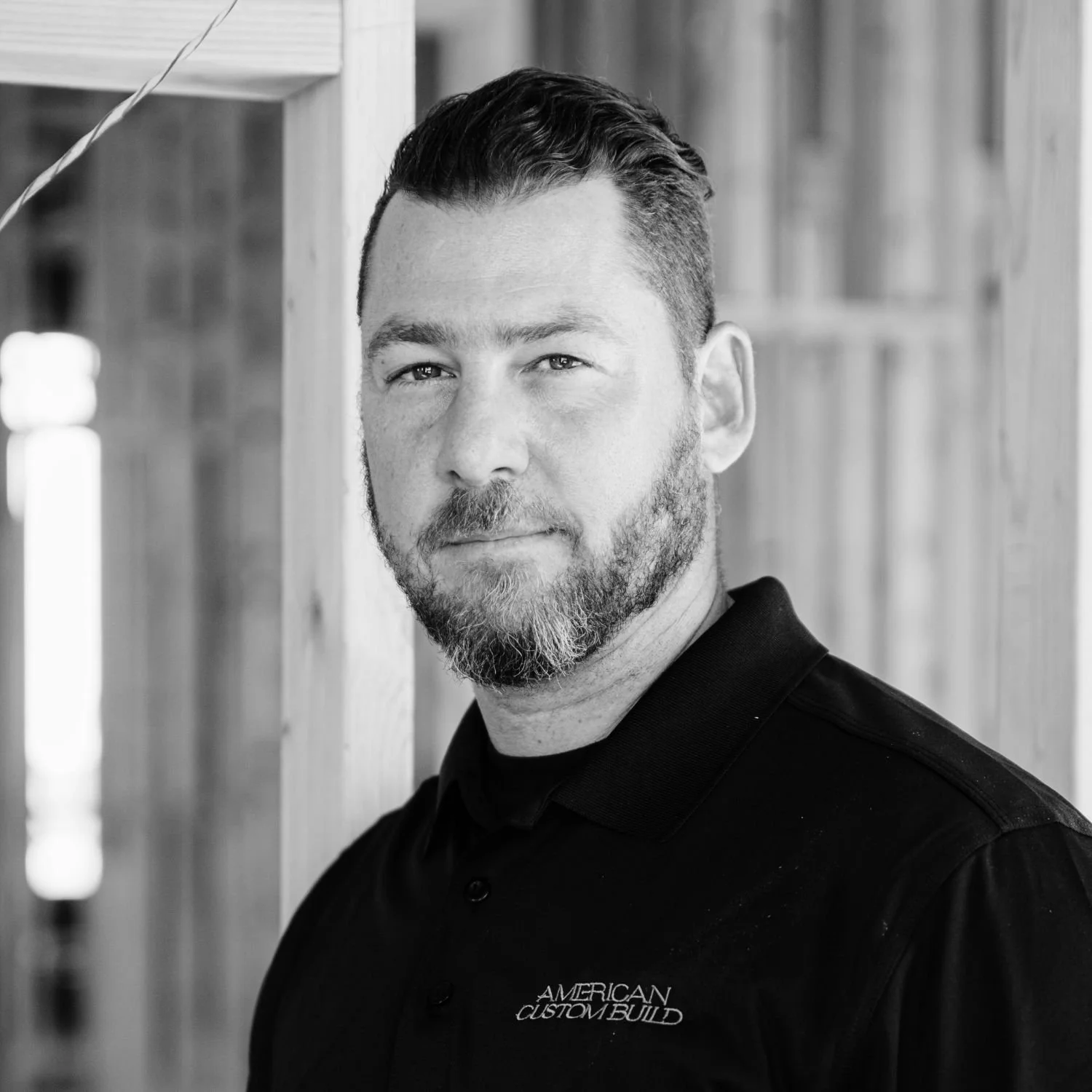 Dimitris, wearing a black polo shirt with a logo that reads 'American Custom Build,' standing in front of a wooden wall.