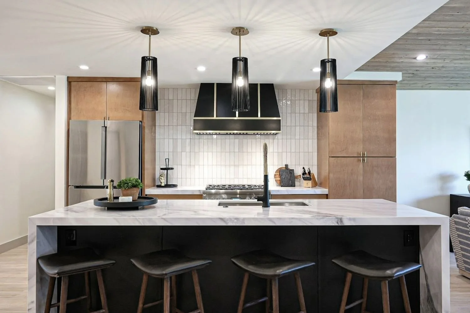 Modern kitchen with a large white marble island, black stools, pendant lights, a stainless steel refrigerator, a black range hood, and wooden cabinets.