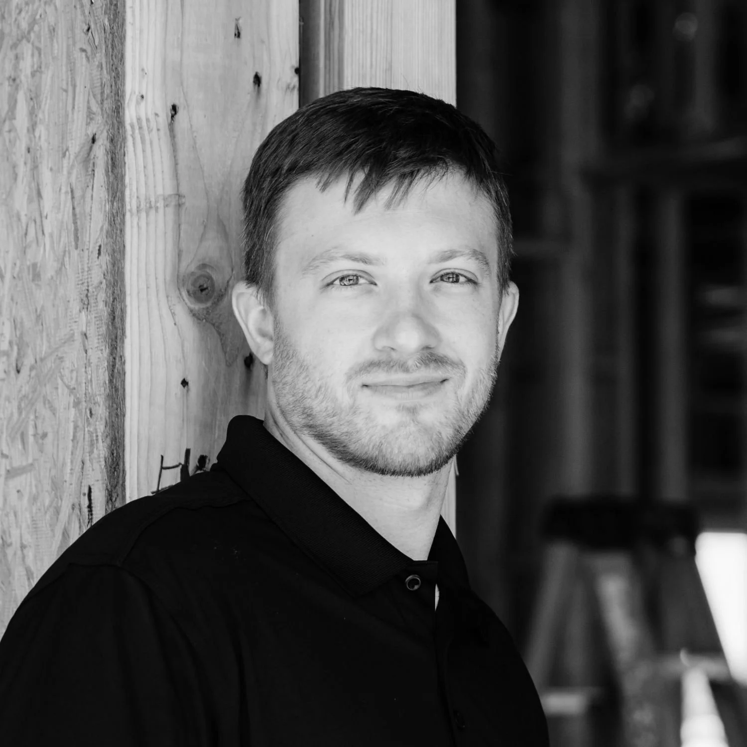 Berkley, wearing a black collared shirt, standing beside a wooden wall and smiling slightly.