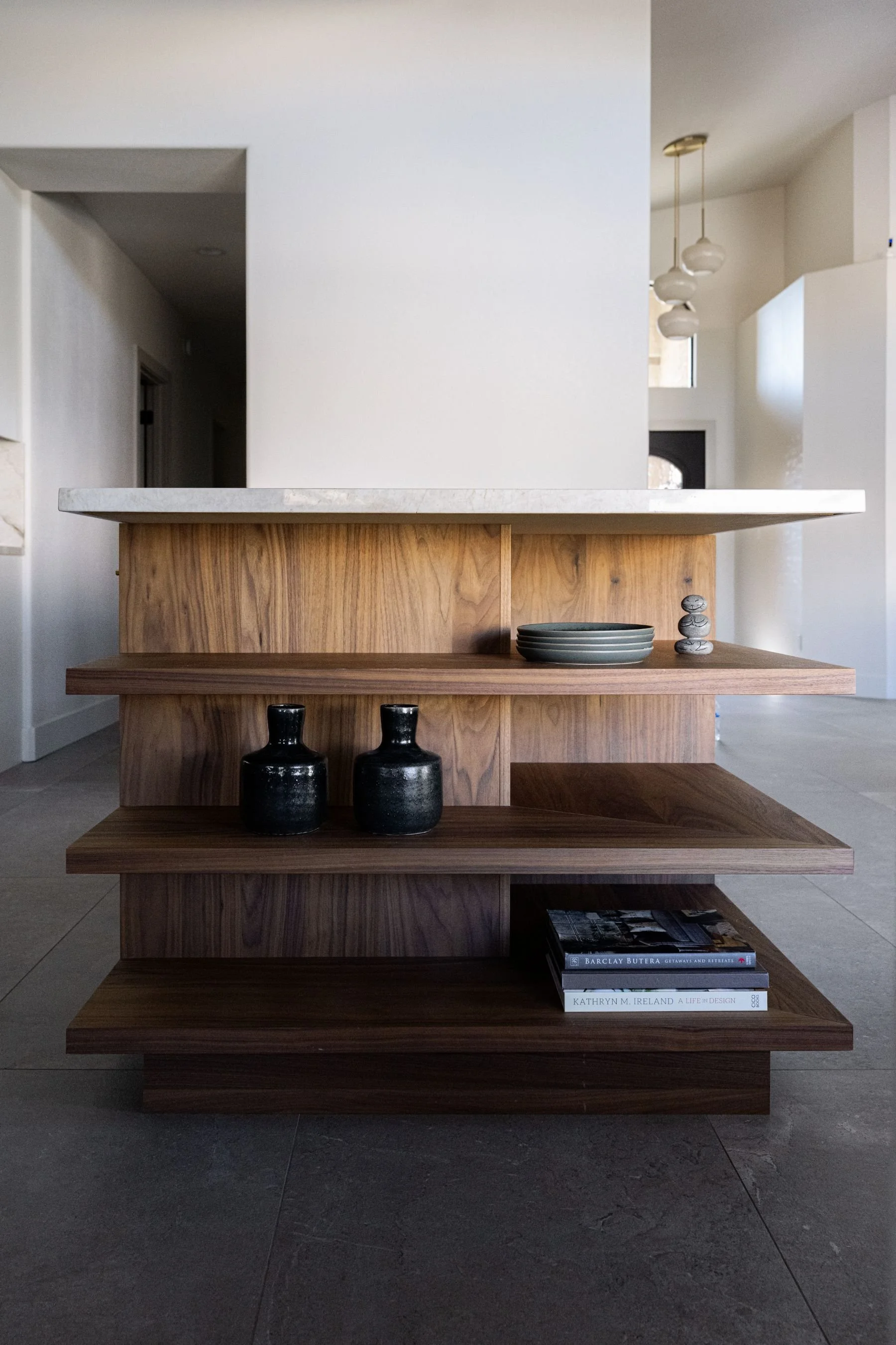 A modern wooden open shelving unit with three shelves, decorated with black vases, ceramic bowls, a stack of smooth stones, and books, in an interior space with white walls and contemporary lighting.