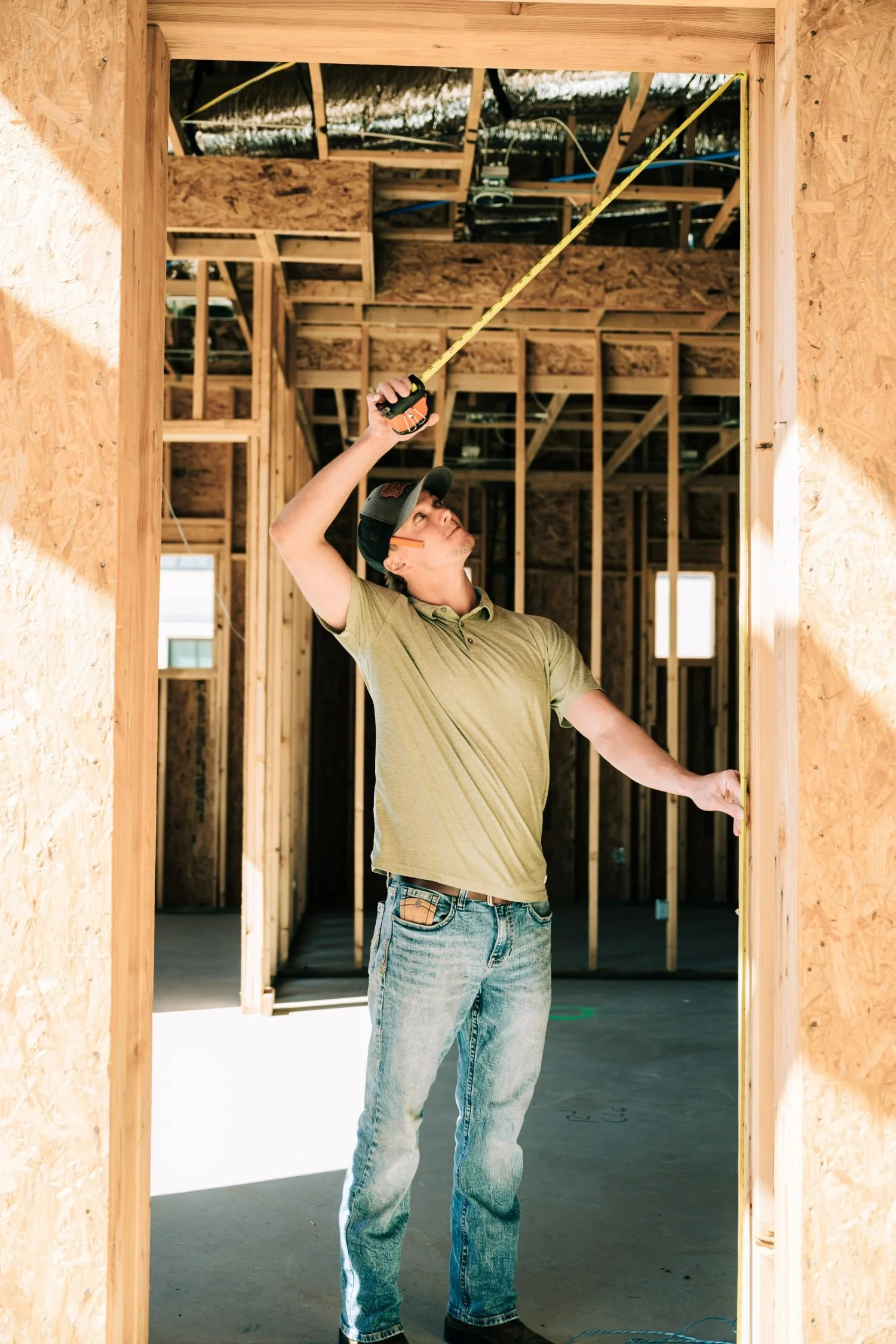 A man in a yellow-green polo shirt and jeans measures a doorway frame inside a wooden house under construction, using a tape measure.