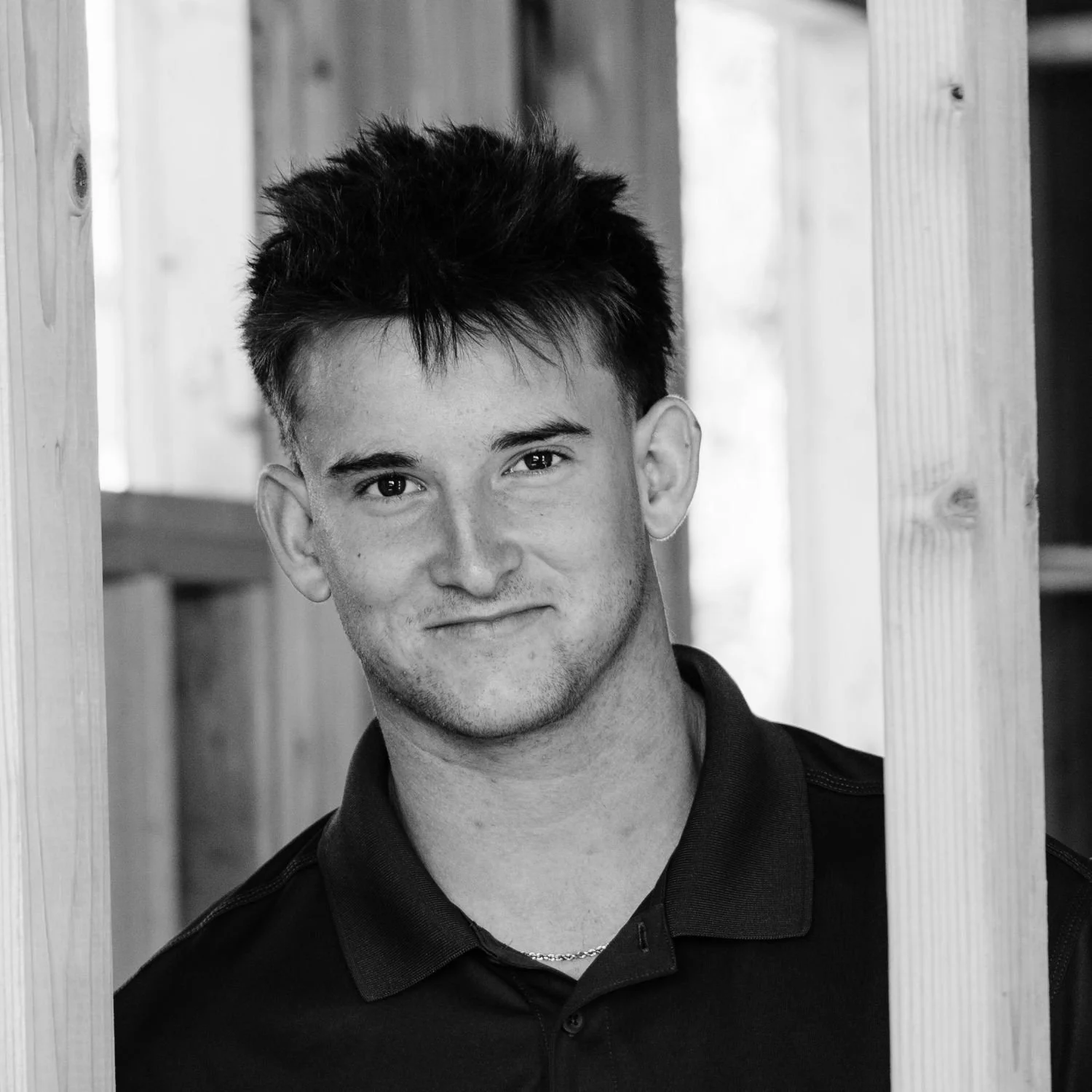 Travis, smiling softly, wearing a dark collared shirt, standing in front of wooden beams.