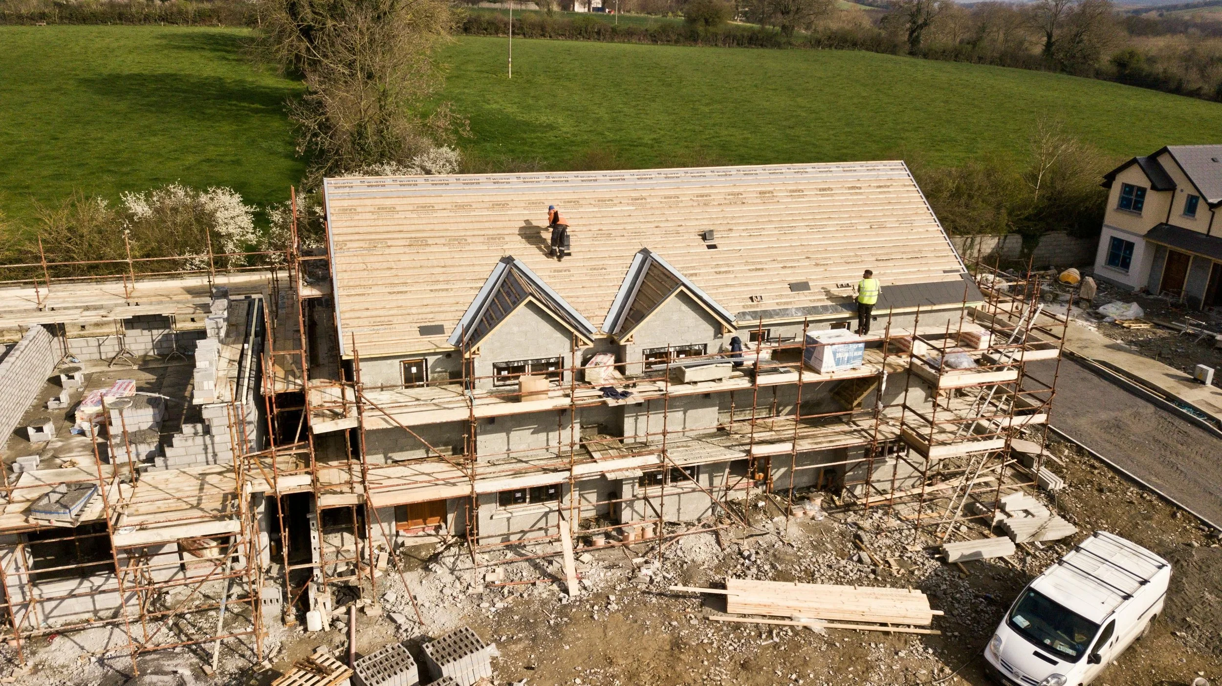 An aerial view of a residential building under construction, with workers on the roof and scaffolding around the structure, and green fields in the background.