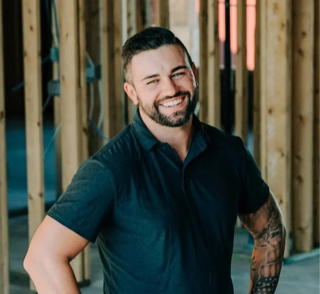 John smiling and standing in a building under construction with wooden framing in the background.