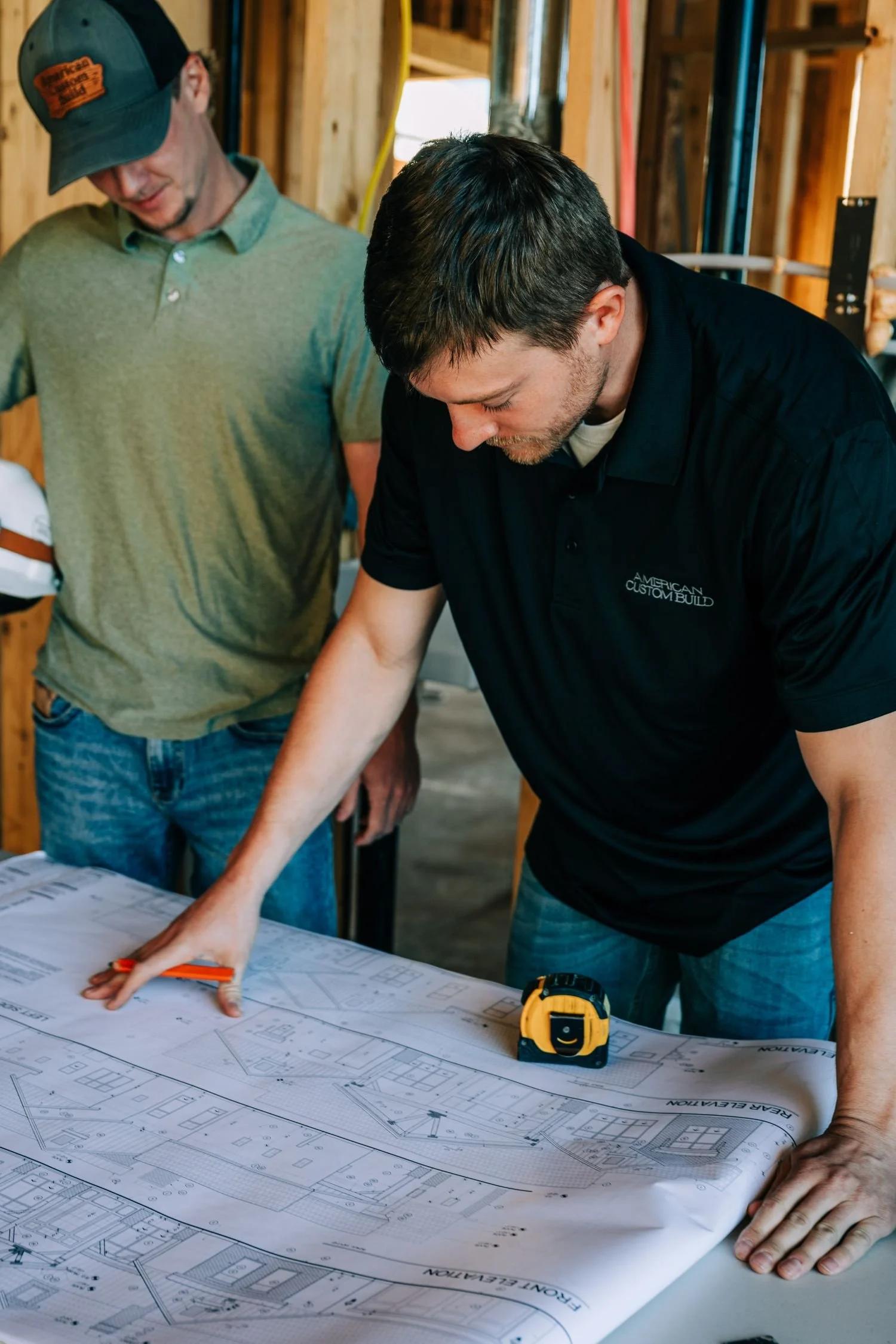 Two men examining architectural blueprints on a table inside a construction site, one pointing at the plans with a pencil and the other observing.