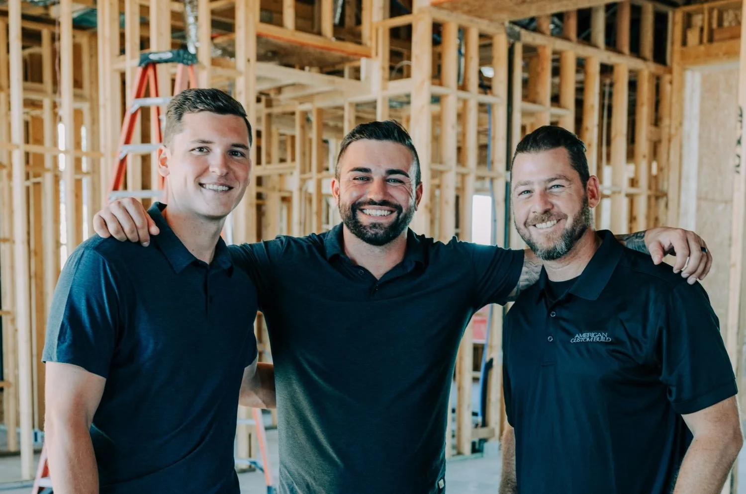 Three smiling men standing arm-in-arm inside a wooden frame structure under construction.