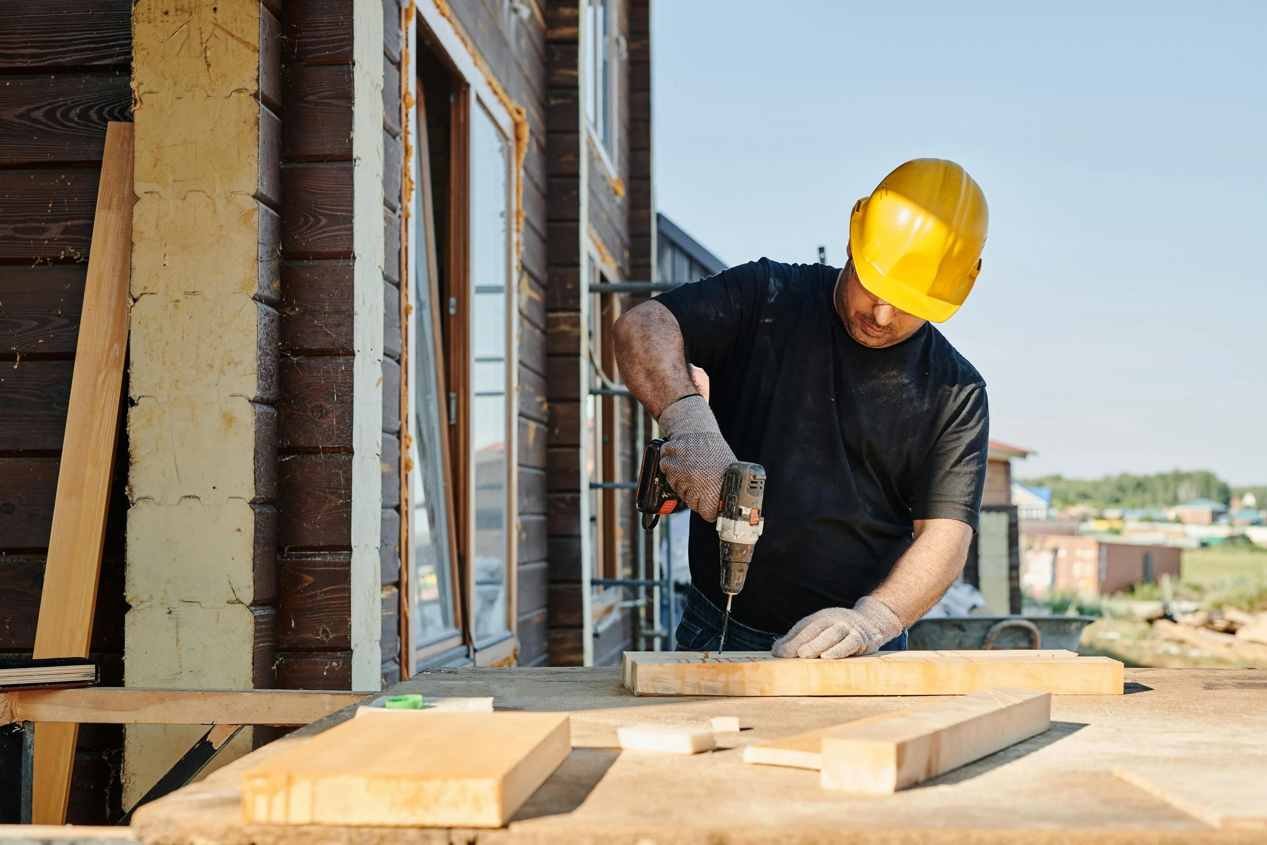 A construction worker wearing a yellow hard hat and black shirt using a cordless drill on a piece of wood at a construction site.