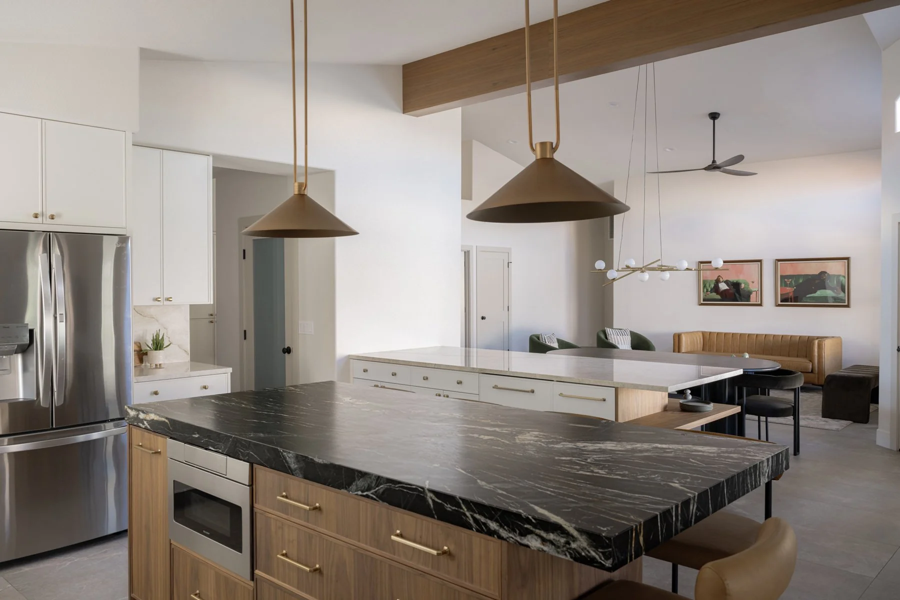 Open kitchen with black marble island on walnut cabinets, brass cone pendants, white walls, and wood beam leading to the living room