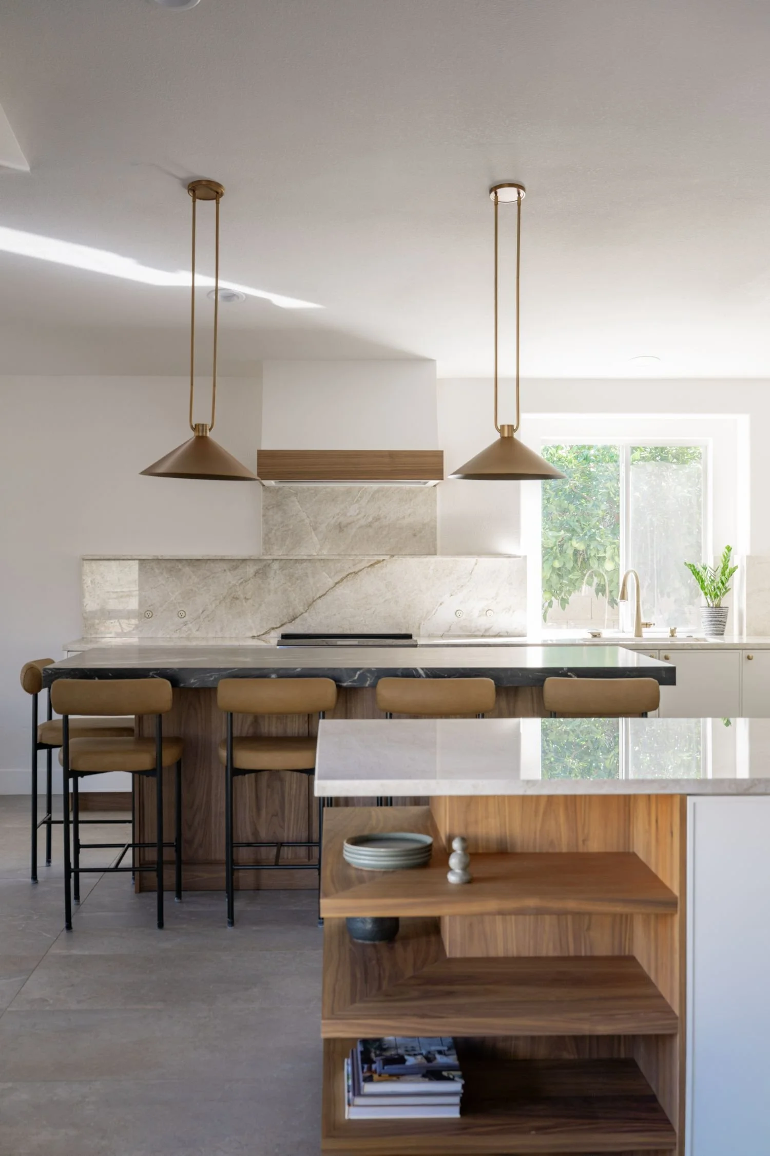 Modern kitchen with a marble backsplash, black island with beige barstools, pendant lights, a window with greenery outside, and wooden open shelves with decorative items.