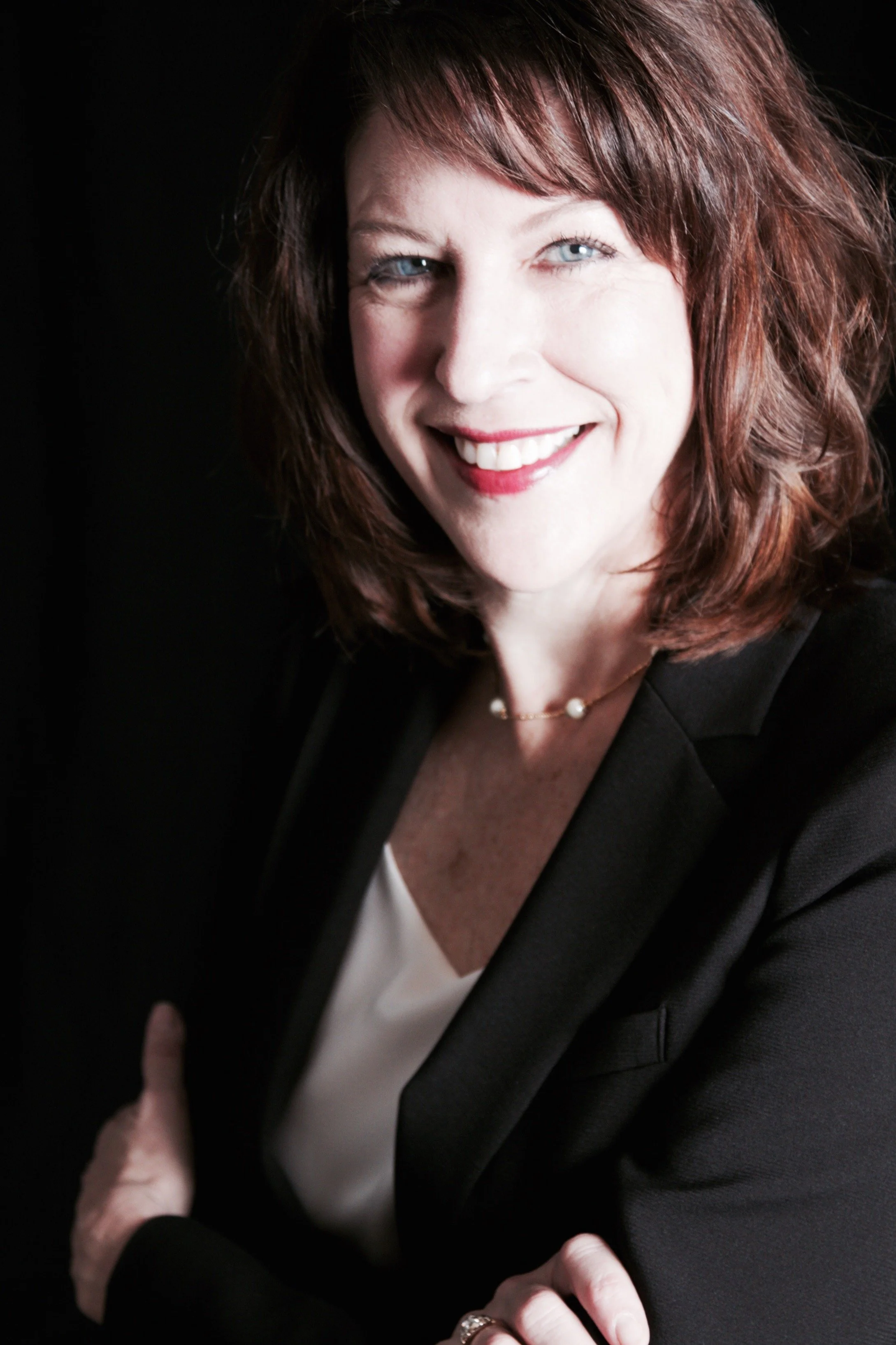 A woman with shoulder-length red hair, blue eyes, and fair skin, smiling, wearing a black blazer, a white blouse, a pearl necklace, and a ring, posed against a dark background.
