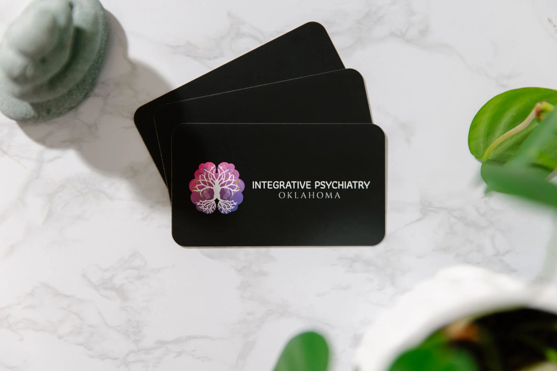 Black business cards with a colorful tree logo and the text "Integrative Psychiatry Oklahoma" on a white marble surface, surrounded by green plants and a small decorative object.