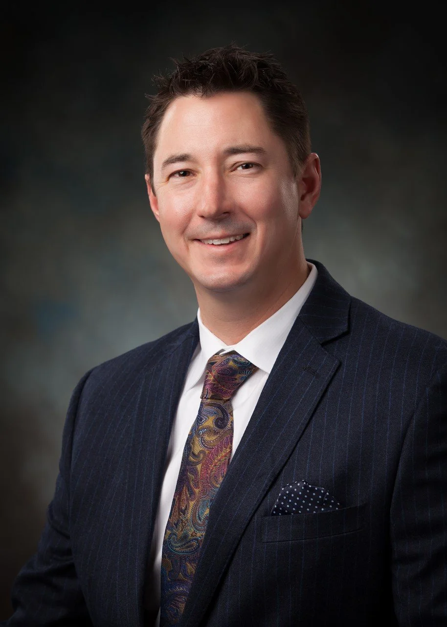 A professional man smiling, wearing a dark pinstripe suit, white shirt, patterned tie, and pocket square, against a dark gradient background.