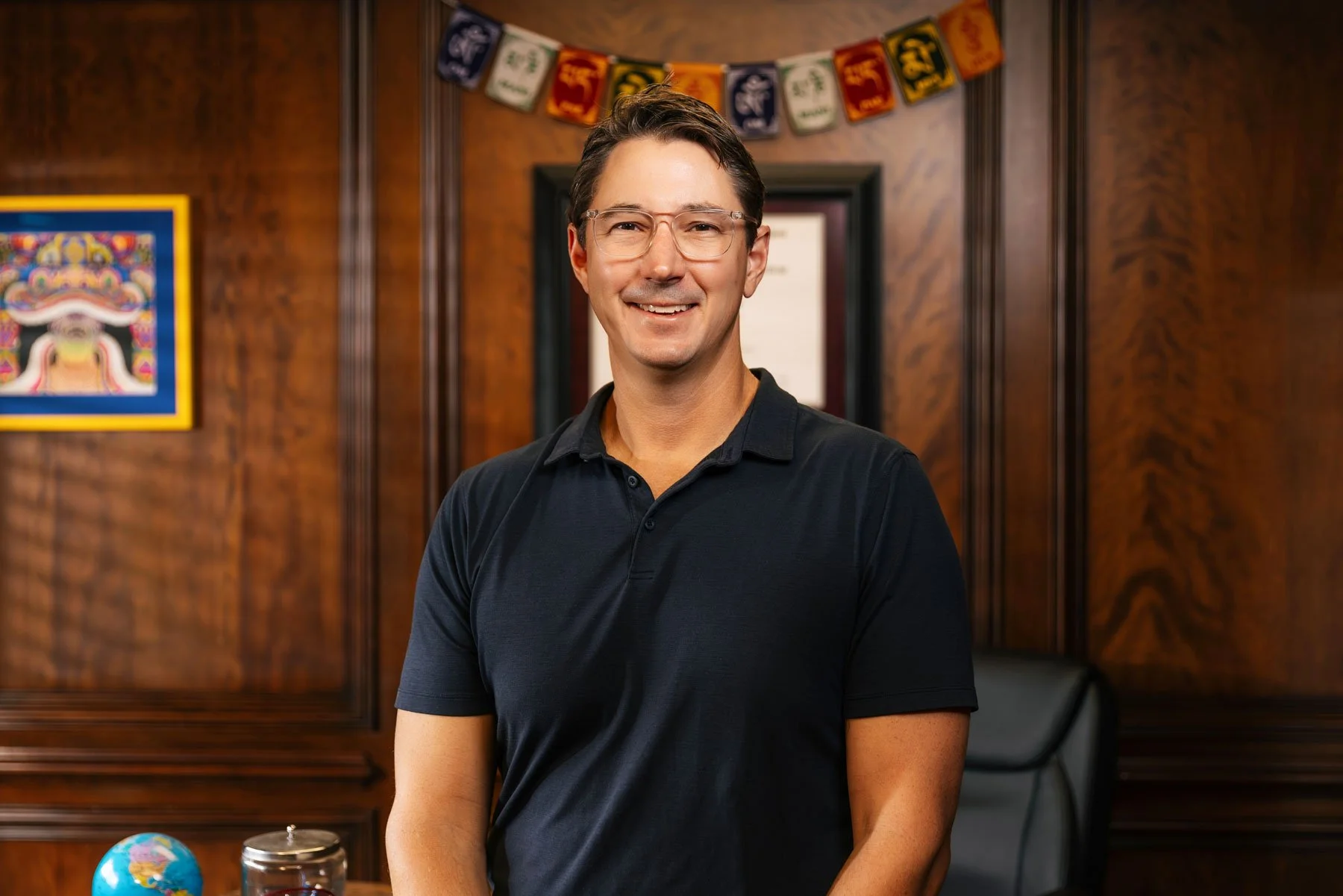 Man in dark polo shirt smiling in office with wood-paneled walls, colorful banner, framed artwork, and small globe.