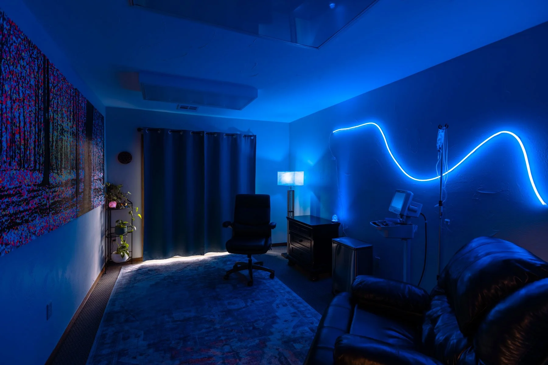 A dimly lit room with blue neon lighting on the wall, a black office chair in front of a window with dark curtains, a small side table with a lamp, a leather couch, a bookshelf with plants, and colorful artwork on the opposite wall.