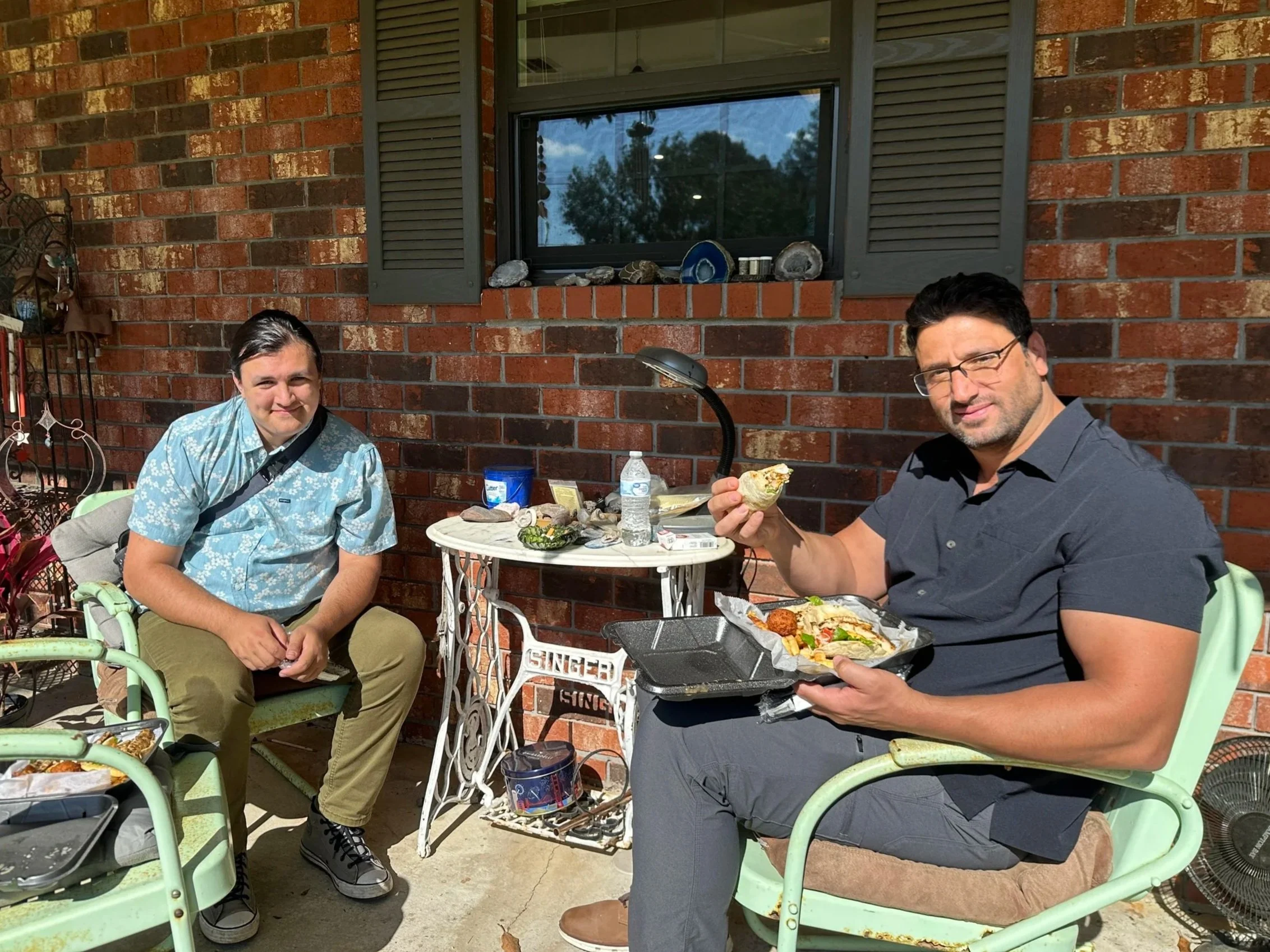 Two men sitting outside at a table with food, in front of a brick wall with a window. One man is holding a slice of pizza, and the other is sitting in a green chair.